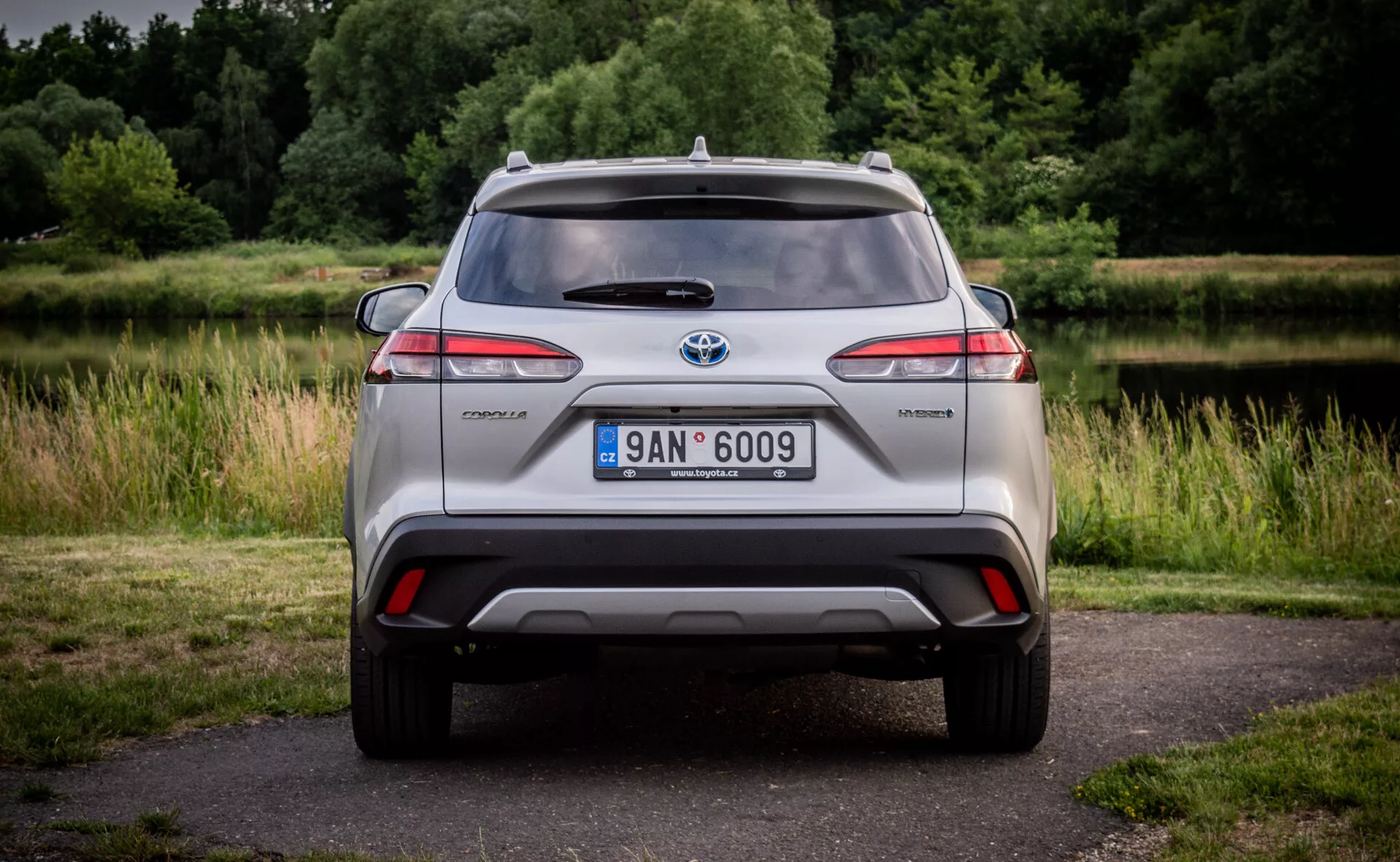 Baksida av en Toyota C-HR hybridmodell som visar LED bakljus, integrerad diffusorstötfångare med reflektorer, takmonterad antenn och lättmetallhjul, mot en naturlig bakgrund