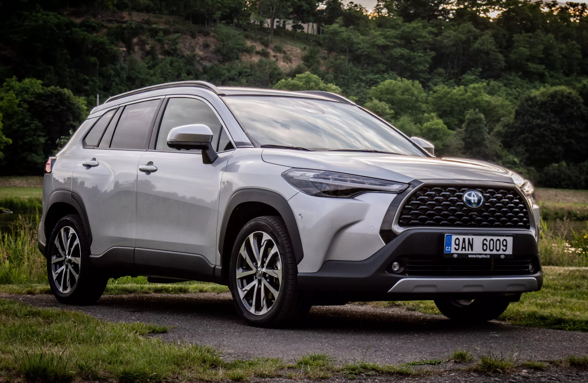 Toyota Corolla Cross 2022 i glansig metallicfinish som visar sin moderna SUV-design med detaljerad frontgrill, emblem, vinklade LED-strålkastare, aerodynamisk frontstötfångare, konturerad motorhuv, sidoprofil med flerstråliga legeringshjul och takräcken, tagen från en låg vinkel för att betona dess höjd och hållning under dagtid i en naturlig förorts miljö.