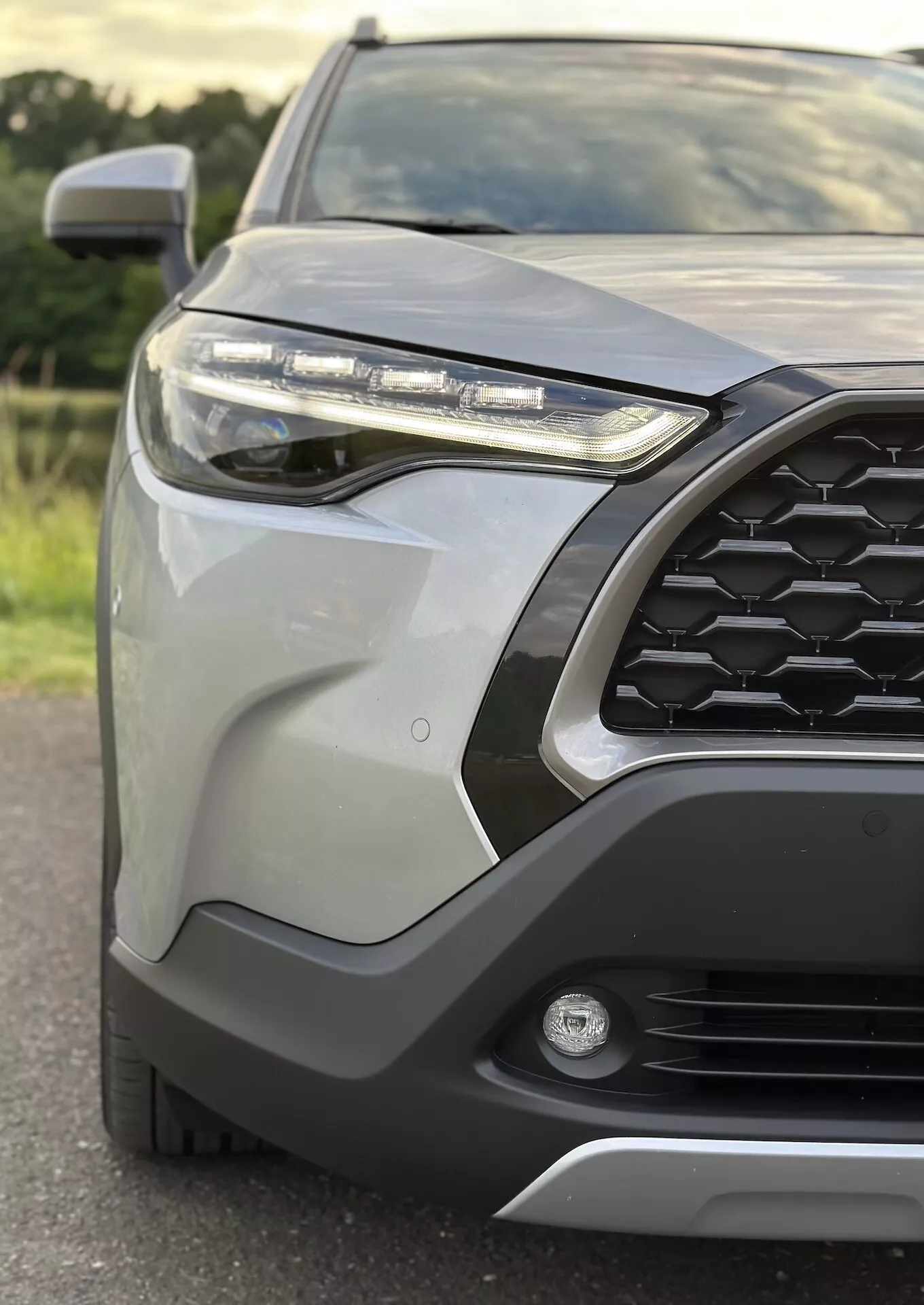Närbild av främre vänstra kvarten av Toyota Corolla Cross 2022 som framhäver hexagonalt grillmönster, LED-strålkastare, dimljus, främre stötfångare med skyddande klädsel, eleganta karosspaneler, integrerad blinkers i sidoupputspeglar, och metallisk finish