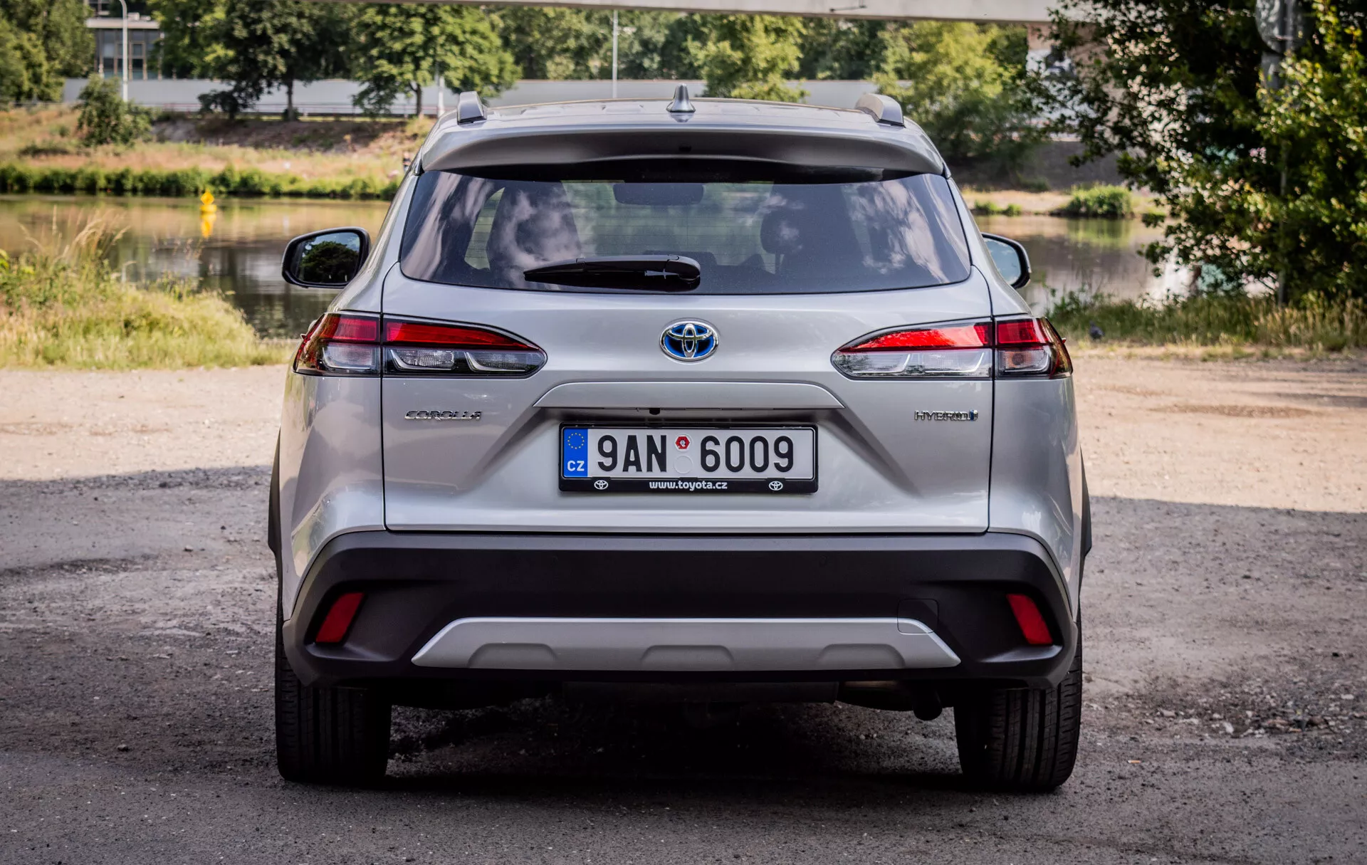 Bakvy av Toyota Corolla Cross 2022, som visar LED-bakljus, hybridmärke och kromad Toyota-emblem med naturligt dagsljus och en naturskön bakgrund av grönska och vatten