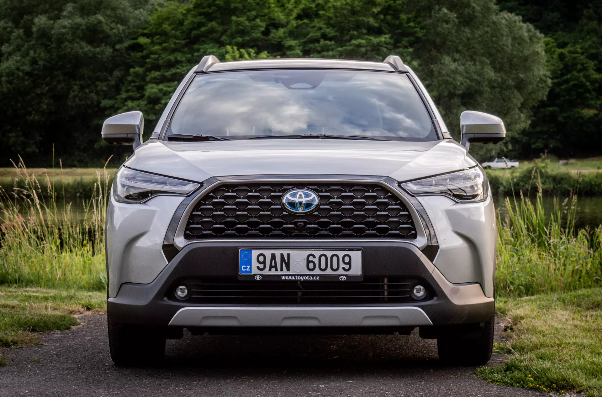 Framsida av en Toyota Corolla Cross 2022, med fokus på dess stora grill med emblem, moderna LED strålkastare, dimljus, registreringsskylts placering, integrerade parkeringssensorer eller framåtriktad kamera, aerodynamiska motorhuvskonturer, grov design på nedre stötfångare och luftintag, parkerad på asfalt med en naturlig bakgrund