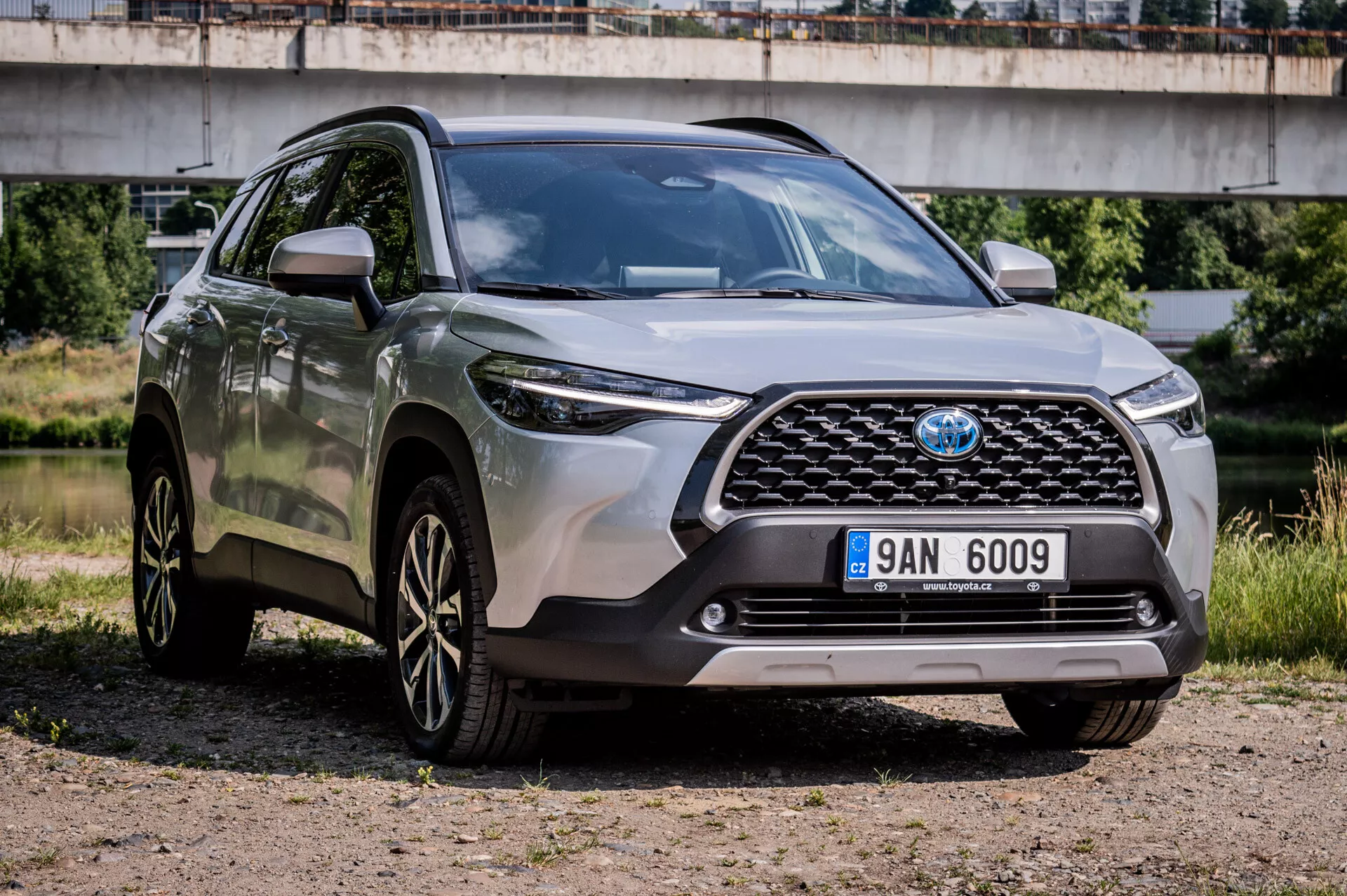 Toyota Corolla Cross 2022 parkerad på grus som visar sin robusta design, sportiga flerspakiga lättmetallfälgar, avancerade LED-strålkastare och hög markfrigång i en tre kvarts främre vy som framhäver frontgrillen, stötfångaren och förarsidans egenskaper med en bro i bakgrunden i en halvurban miljö