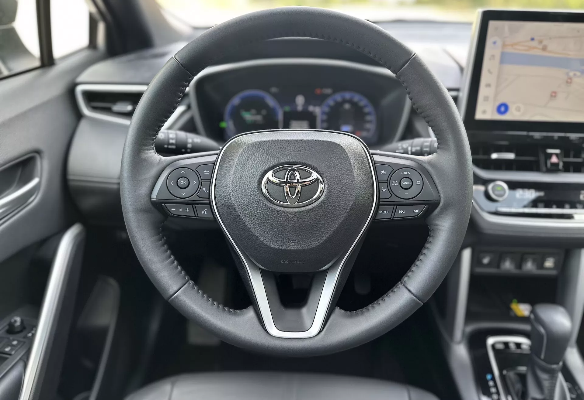 Interiör av Toyota Corolla Cross 2022 som visar ratten med Toyota-logotypen, integrerade kontroller för ljud, samtal och farthållare, bakom vilken en digital instrumentbräda med hastighetsmätare, varvräknare och multifunktionell display delvis syns, inklusive en större central instrumentbräddisplay för GPS-navigering, allt mot en bakgrund av samtida mörkt materialdesign.