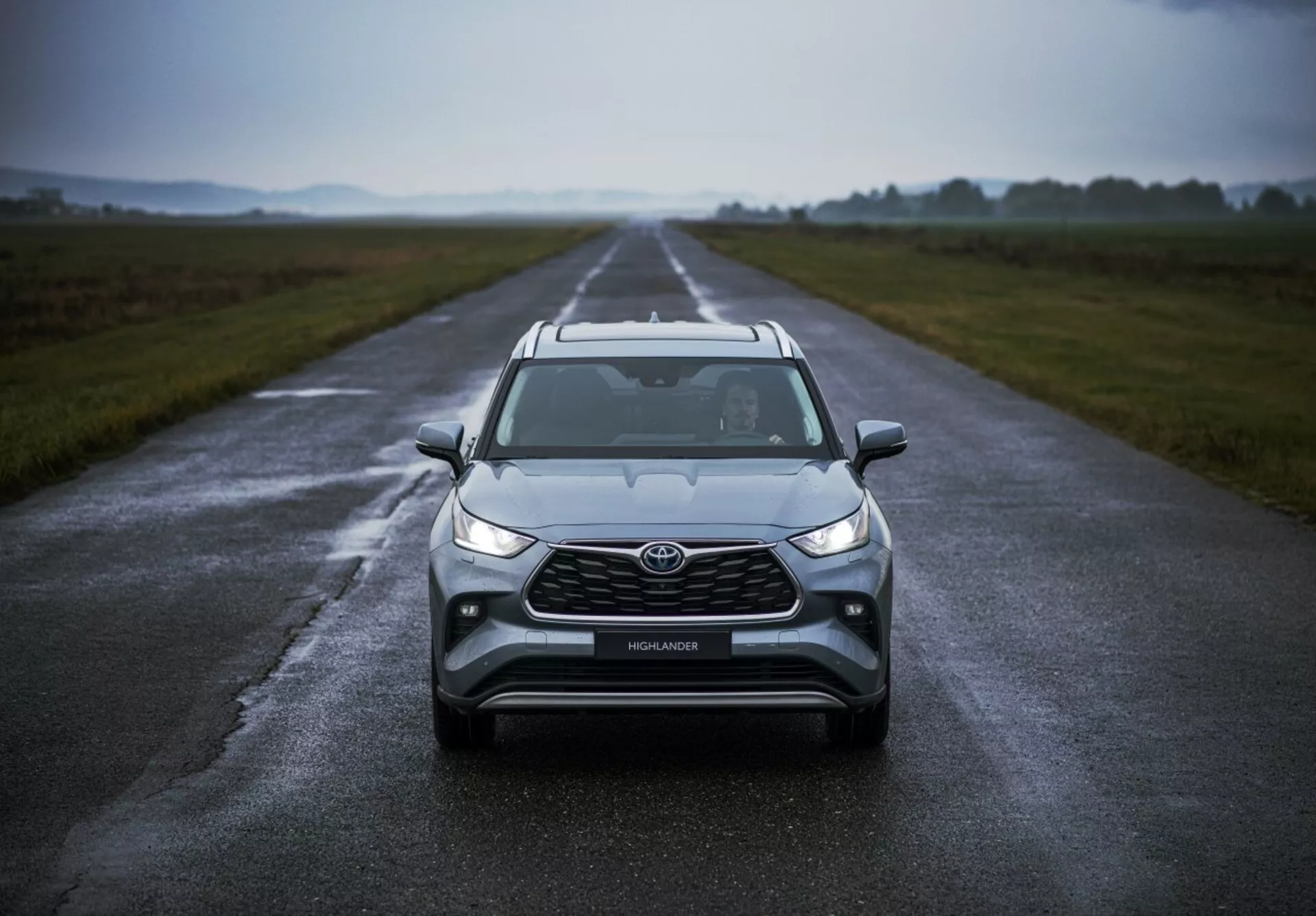 Toyota Highlander 2021 placerad på en våt väg som framhäver dess frontdesign, molnigt väder, med LED-strålkastare, dimljus, en stor grill och avancerad säkerhetsteknik