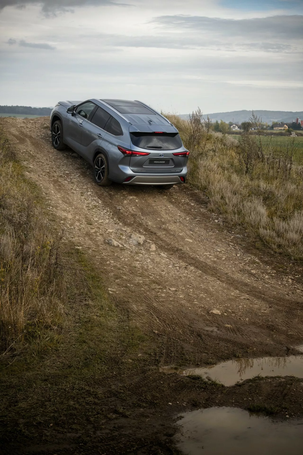 2021 Toyota Highlander mörk halvkombi fångad från en hög vinkel på en grusväg, visar baksidan tre fjärdedels vy, sportig design och potential för lätt terrängfärd mitt i ett lantligt landskap