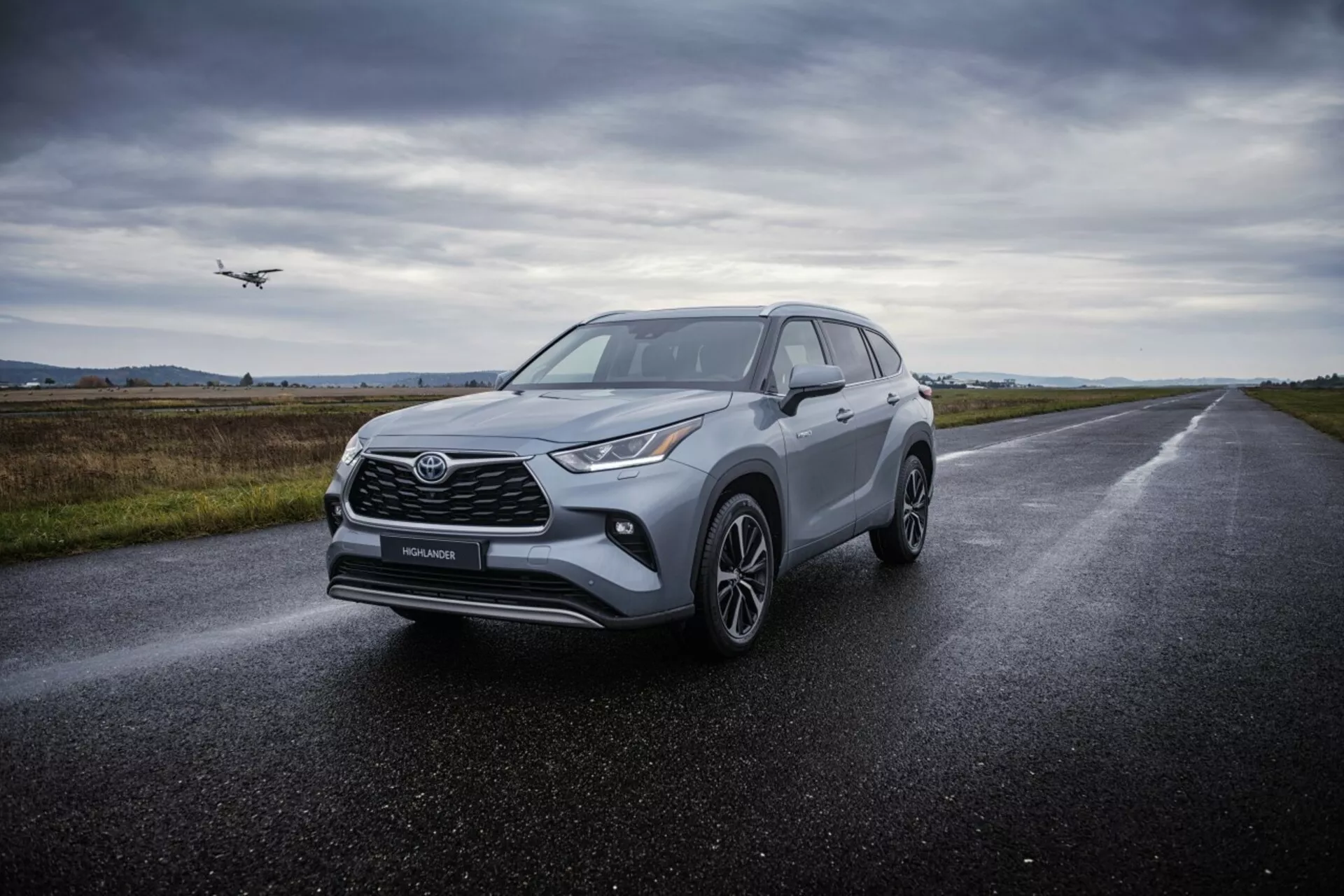 Crossover SUV Toyota Highlander 2021 på våt asfaltväg, visar dess vinklade strålkastare, framträdande frontgrill, flerspåriga lättmetallhjul och aerodynamisk design under diffust molnigt ljus