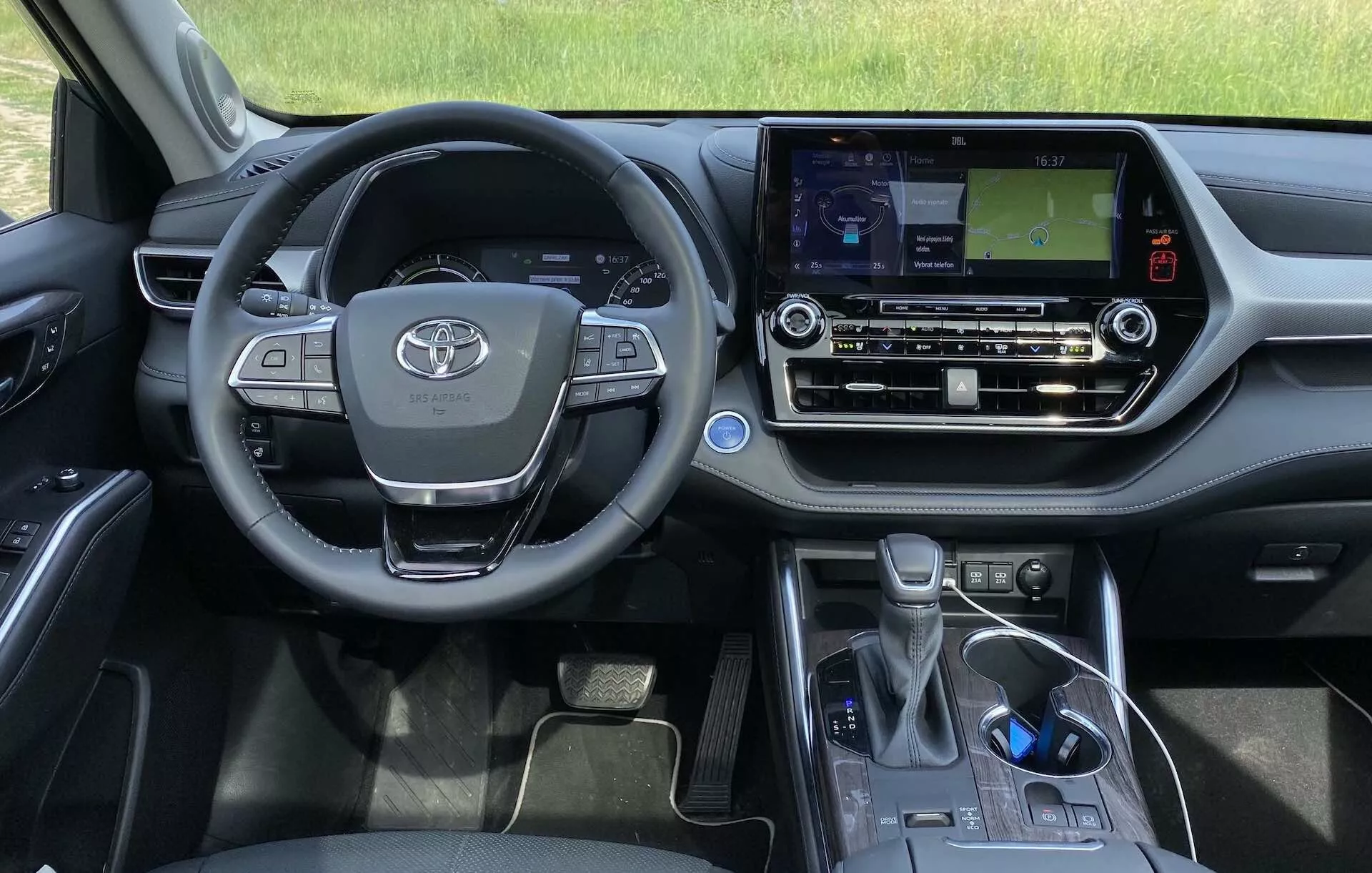 Interiören i 2021 års Toyota Highlander 2.5 Hybrid Business Edition från förarens synvinkel. Bilden visar ratten med Toyota-emblemet, multifunktionella knappar, delvis synlig instrumentpanel och mittkonsol med en pekskärmsinfotainmentsystem som visar en navigationskarta. Klimatkontroller, ett förvaringsfack, den elektroniska växelspaken och parkeringsbromsen är också synliga, vilket visar bilens moderna design och tekniska funktioner.