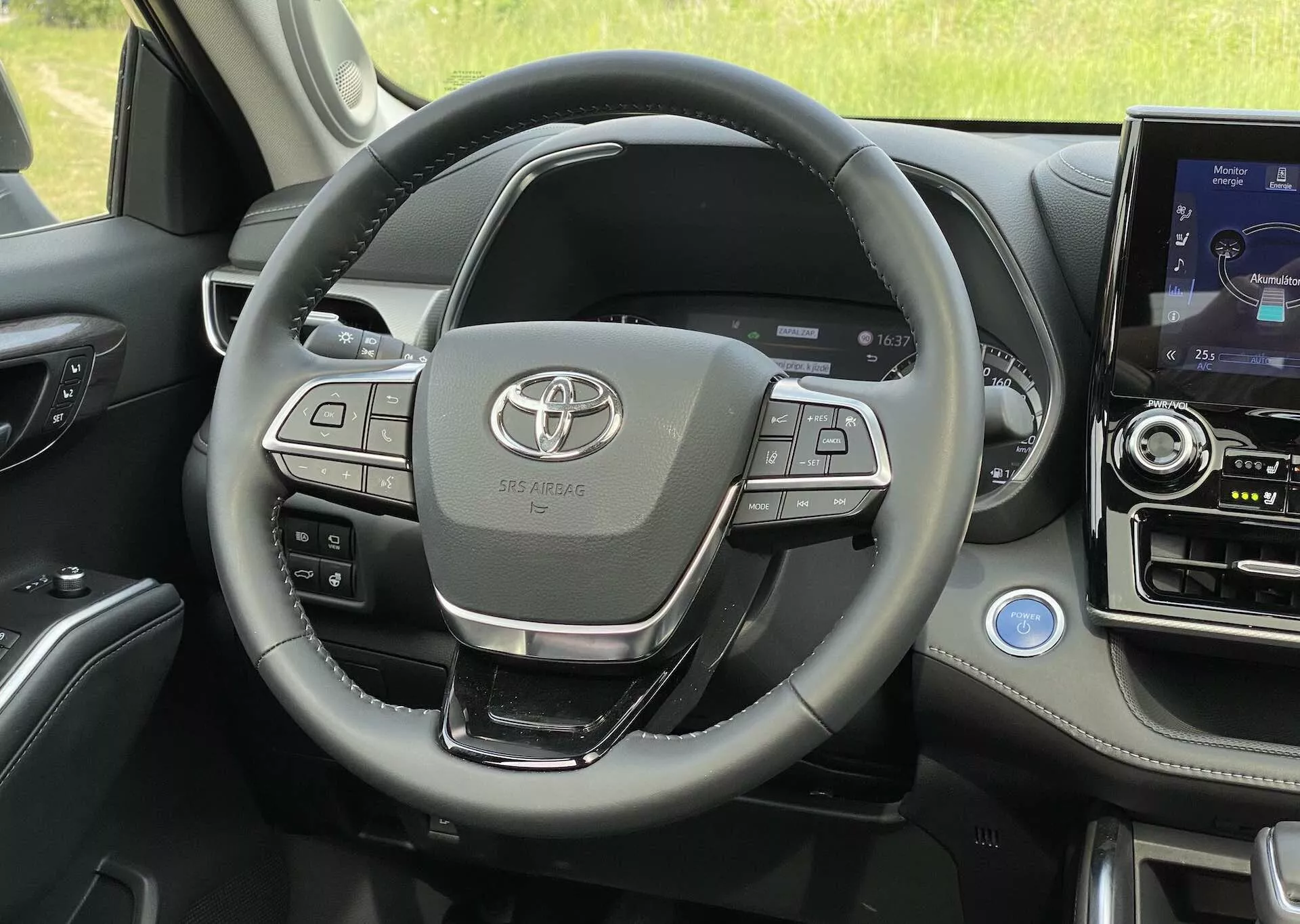 Interiörvy av 2021 Toyota Highlander 2.5 Hybrid Business Edition från en förstapersonsperspektiv, med fokus på den multifunktionella ratten med ljud-, telefonanslutnings- och farthållarknappar, Toyota-emblemet tydligt synligt i mitten. Synliga är också instrumentpanelen, mittkonsolen med infotainmentsystem med pekskärm, växelspaken, dörrkontrollpanelen och luftventilationen, som visar material av hög kvalitet och avancerade funktioner.