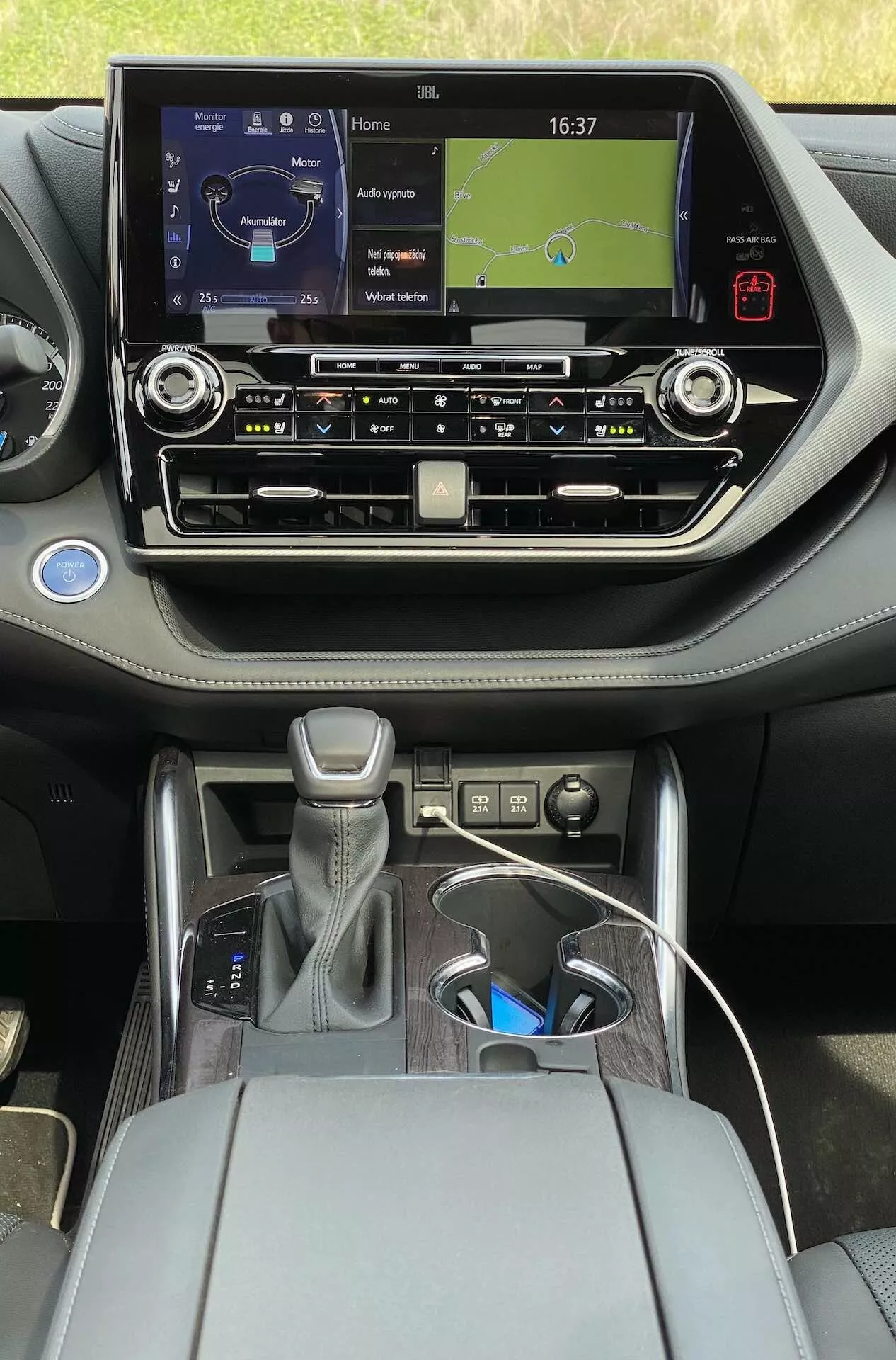 Interiörvy av 2021 Toyota Highlander 2.5 Hybrid Business Edition från baksätet, med ett modernt pekskärmsbaserat infotainmentsystem som visar en klocka och navigationskarta, fysiska knappar för klimatanläggning, luftventiler, en automatisk växelspak med läderskydd och silverdetaljer, en kontrollratt, ett centralt armstöd med öppen förvaringsutrymme, två mugghållare, USB-laddningskabel och en modern tvåtonad design med metalliska accenter.