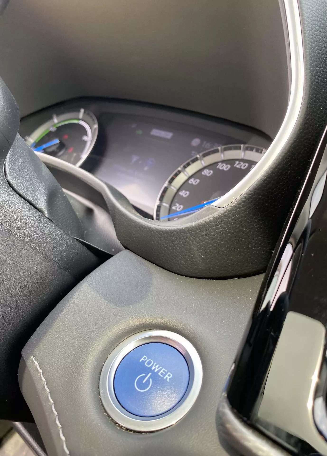 Interiörvy av Toyota Highlander 2021 2.5 Hybrid Business Edition fokuserad på förarens perspektiv. Bilden visar en del av ratten och den digitala instrumentpanelen genom den övre delen av ratten. Panelen innehåller en hastighetsmätare markerad upp till 220 km/h och en energianvändningsmätare, vilket tyder på fordonets hybridkaraktär. En framträdande blå 'POWER'-knapp syns på rattstången, vilket betonar knapplay-systemet. Interiören har moderna svarta plaster och en ratt av läder eller liknande material, med naturliga ljusförhållanden som betonar bilens nya eller väl underhållna skick.