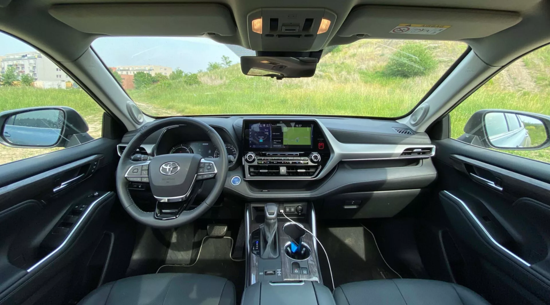 Interiörvy av Toyota Highlander 2021 Hybrid Business Edition från baksätet, som visar en symmetrisk vy av framdaskkonsollen. Ratten, med fordonets emblem och monterade ljudkontroller, syns tydligt till vänster i mitten. Den centrala instrumentpanelen har en infotainment-pekskärm utan fysiska knappar, kompletterad med klimatkontrollknappar och reglage nedanför. Växlaren, en modern elektronisk växelspak, är synlig på den nedre centrala delen av bilden, nära ett eventuellt område för laddning av smartphones. Den lyxiga interiören är markerad av en blandning av lädersäten, mjuka material på instrumentpanelen och kromliknande accenter. Soltaket släpper naturligt ljus in, med kontrollpaneler nära backspegeln. Utanför syns ett naturlandskap genom framsidorutorna.