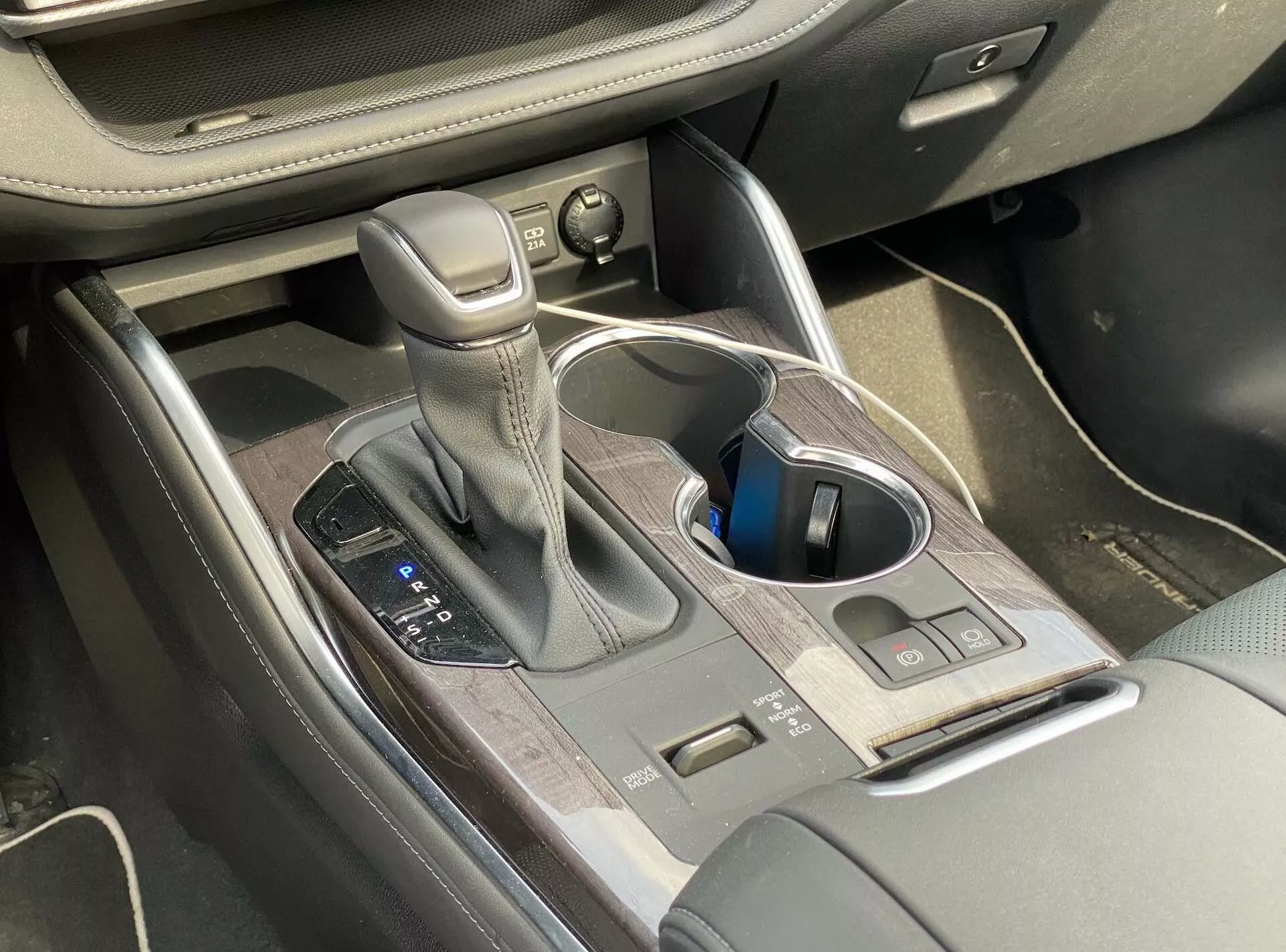 Interiörvy av 2021 års Toyota Highlander 2.5 Hybrid Business Edition med fokus på mittkonsolen, som visar automatväxeln med lädersöm, körlägeskontroller, mugghållare, kontrollknappar, mittkonsolförvaring med träfärdig yta och delvis synliga sätesjusteringskontroller.