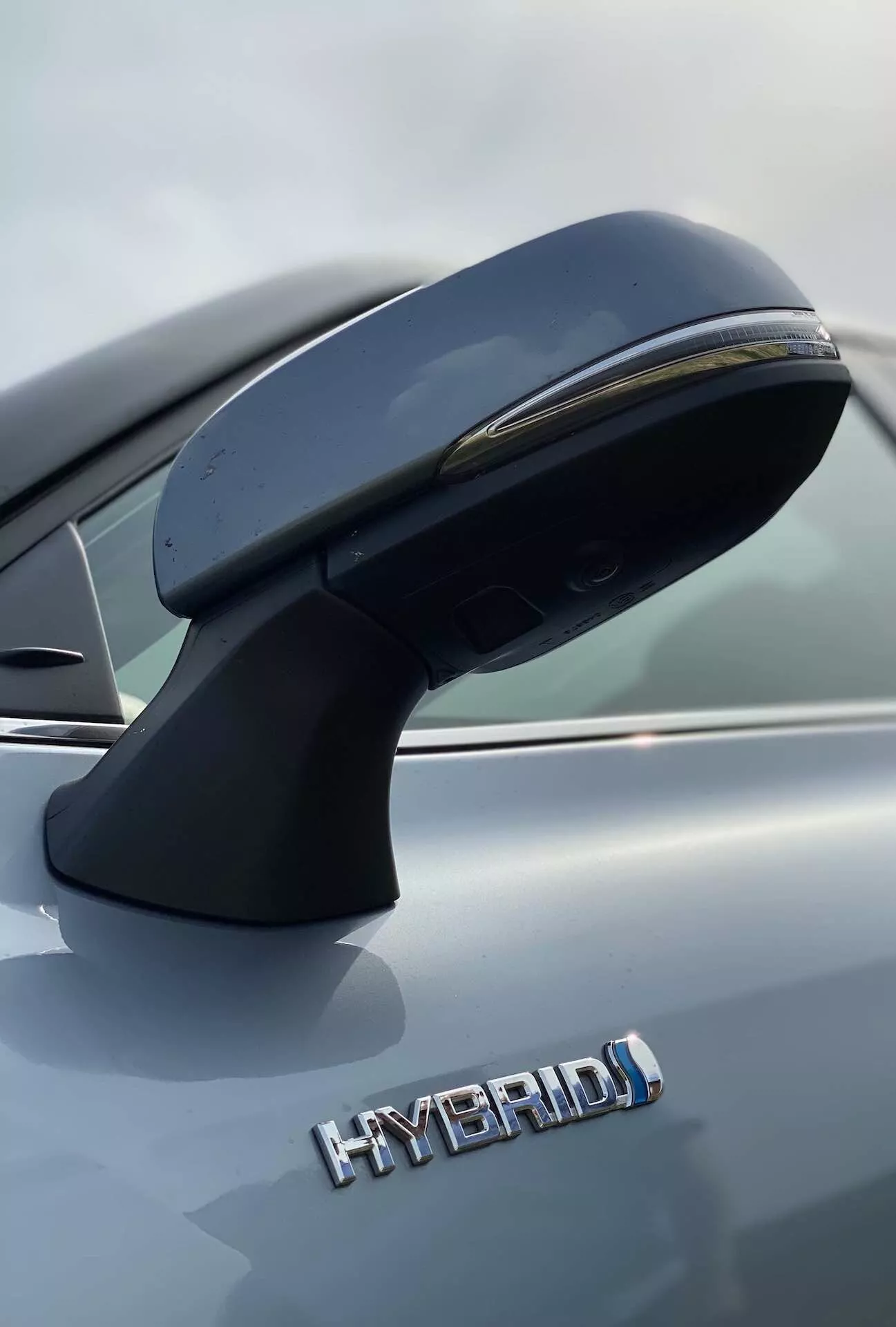 Närbild av förarsidans yttre spegel och delvis sidovy av en Toyota Highlander 2021 Hybrid Business Edition. Spegeln, monterad på dörren, har en strömlinjeformad aerodynamisk design med inbyggd blinkers. Fotot, taget från en låg vinkel, framhäver 'HYBRID'-märket med stora bokstäver som indikerar den miljövänliga hybriddrivlinan. Den metalliska grå finishen på bilen är tydligt synlig. Skarpa detaljer framhävs av det grundskärpedjupet som gör bakgrunden suddig.