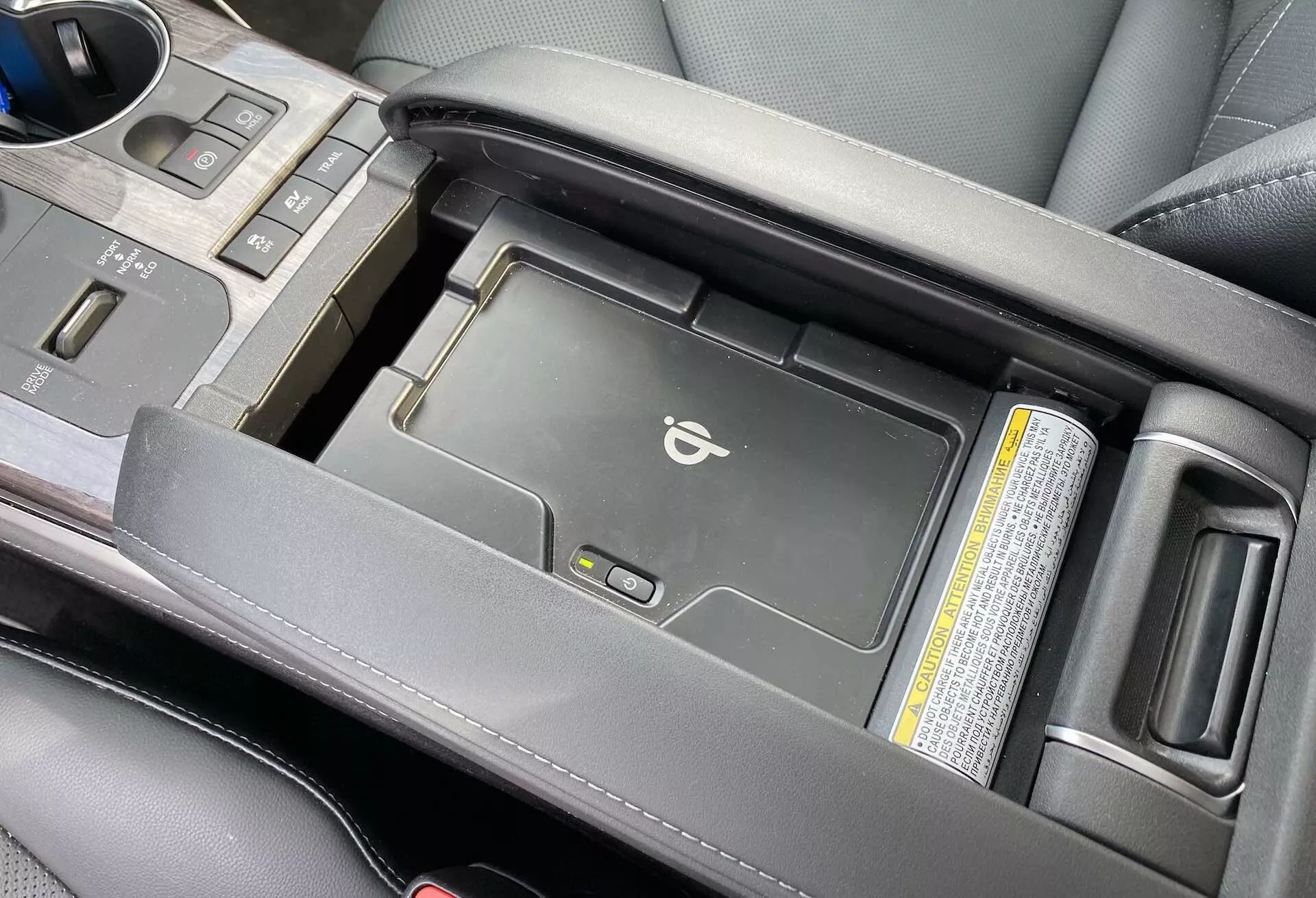 En närbild av mittkonsolen på Toyota Highlander 2021 Hybrid Business Edition från en något upphöjd vinkel, som visar det centrala armstödsområdet delvis öppet för att avslöja en trådlös laddningsplatta för mobila enheter. Bilden framhäver bilens premium svarta läderarmstöd, växelspak och mugghållare, med fokus på moderna tekniska bekvämligheter och estetisk kvalitet.
