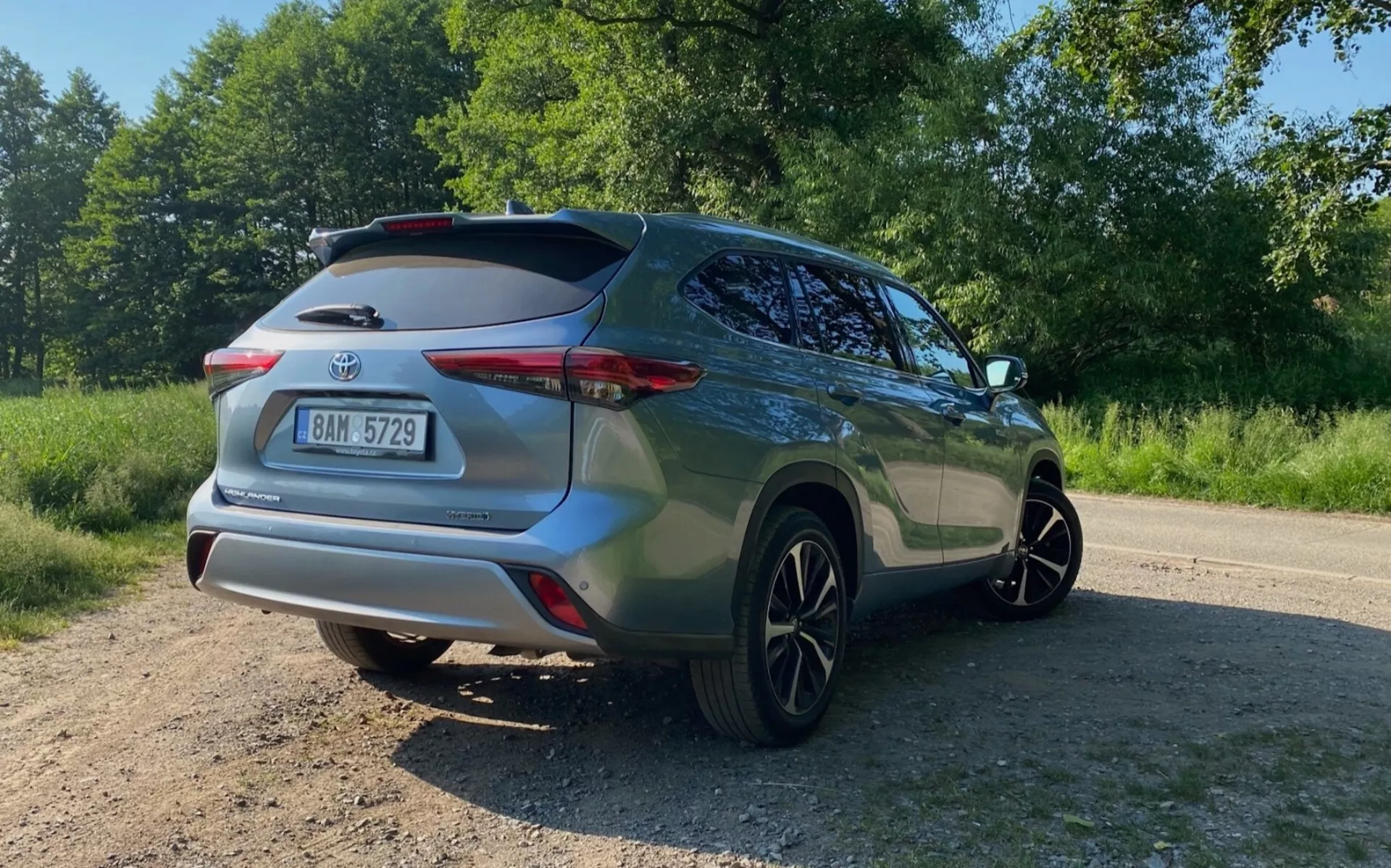 Trekvarts bakifrån vy av en 2021 Toyota Highlander 2.5 Hybrid Business Edition SUV, fångad från marknivå med en uppåtvinklad vy. Bilden betonar den högra baksidan, inklusive bakre stötfångaren, baklyktan, bakluckan med takmonterad spoiler, passagerardörren, fönstret och bakhjulet. SUV:ens metalliska färg glittrar i solljuset, placerad på en ojämn, oasfalterad yta omgiven av grönska.