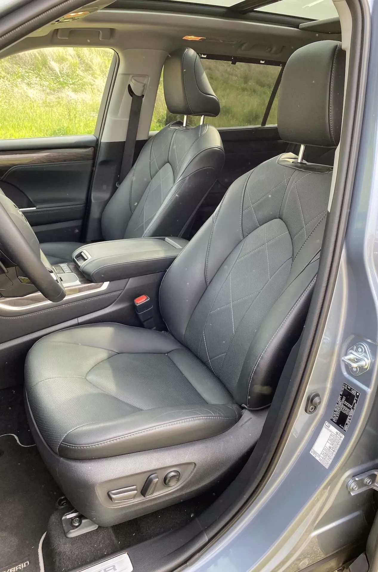 Interiören i Toyota Highlander 2021 2.5 Hybrid Business Edition som visar förarsätet med svart quiltat läderklädsel, sidostöder och eljusteringskontroller. Utsikten inkluderar en del av mittkonsolen samt dörrpanelen med kontroller för elhissar och speglar, högtalargaller och säkerhetsbälte fram. Den lyxiga och ergonomiska designen framhävs av naturligt ljus.