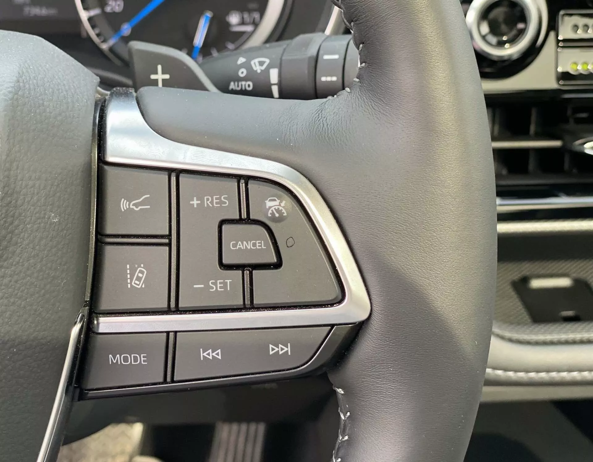 Närbild av vänstra sidan av ratten i en Toyota Highlander 2021 2.5 Hybrid Business Edition, som visar svart läder med synliga sömmar, silver/krom accenter, knappar för adaptiv farthållare och multimedia system, och en del av instrumentpanelen och mittpanelen.