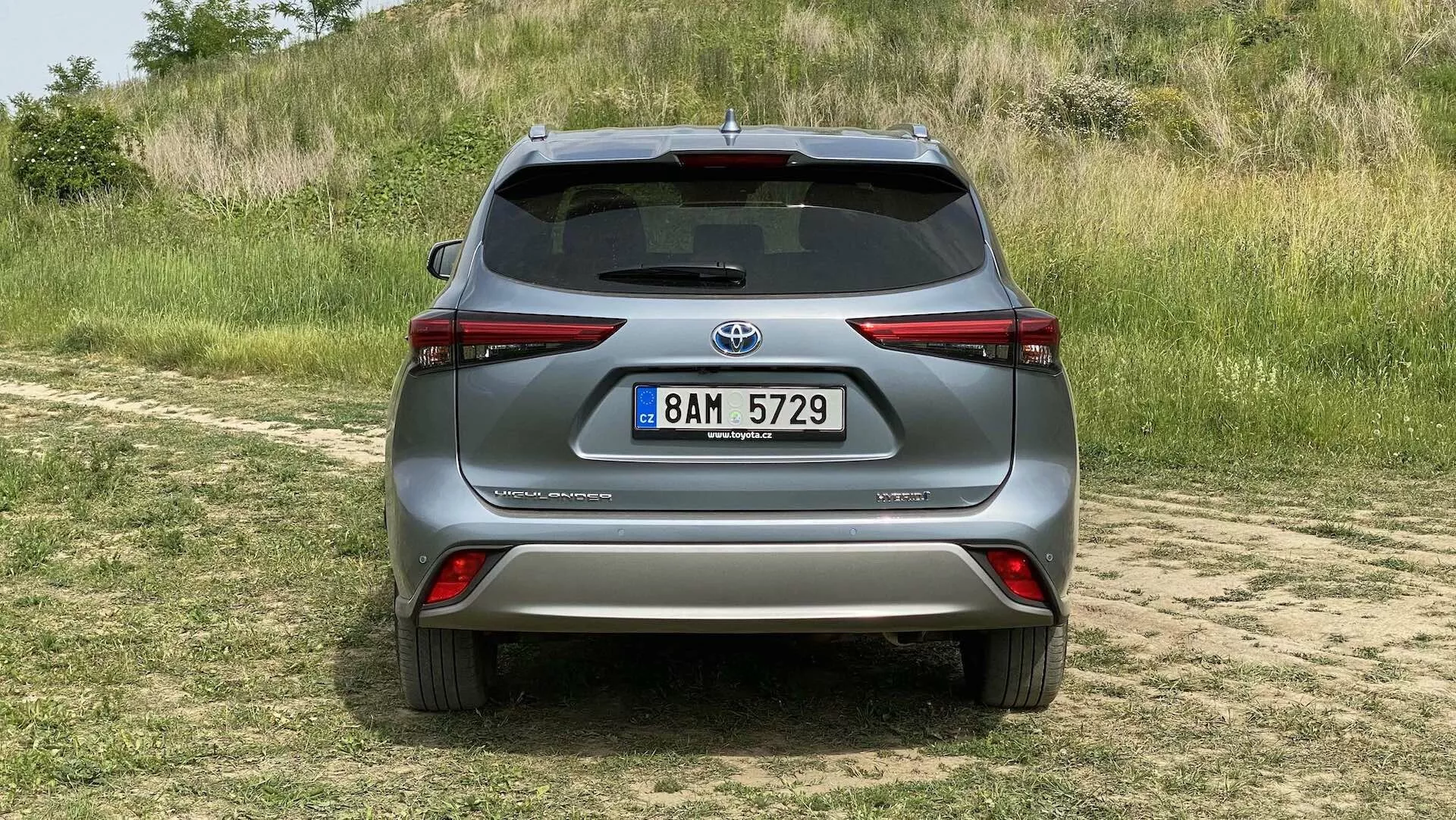 Bakre vy av en silverfärgad Toyota Highlander 2021 2.5 Hybrid Business Edition parkerad på en gräsyta med en dammig stig i förgrunden, fotograferad i dagsljus med naturligt ljus. Den moderna SUV:n har eleganta baklyktor som sveper runt sidorna, en framträdande bakspoiler, integrerade reflexer på stötfångaren, en backkamera ovanför registreringsskylten och en silverfärgad skyddsplåt under stötfångaren, vilket antyder dess terrängkapacitet och teknologiska framsteg i parkeringsassistans och backspegelsystem.