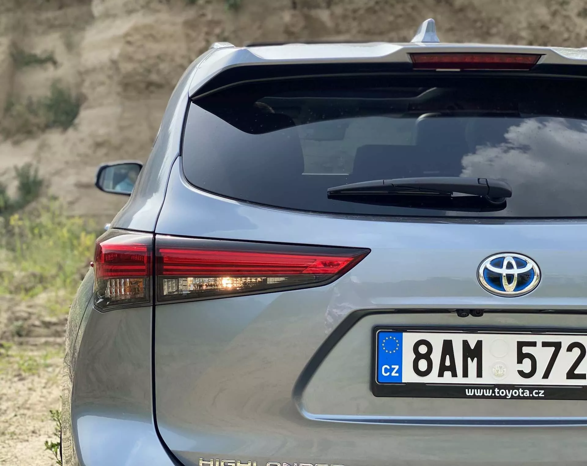 Närbild av bakre delen av en Toyota Highlander 2021 Hybrid Business Edition i silver, fotograferat från en trekvartsvinkel som framhäver höger sida. Innehåller ett distinkt LED-bakljus, europeisk stils registreringsskylt från Tjeckien, hybridemblem, bakrutedfrostersystem och högt monterat bromsljus mot en naturlig, jordnära bakgrund.
