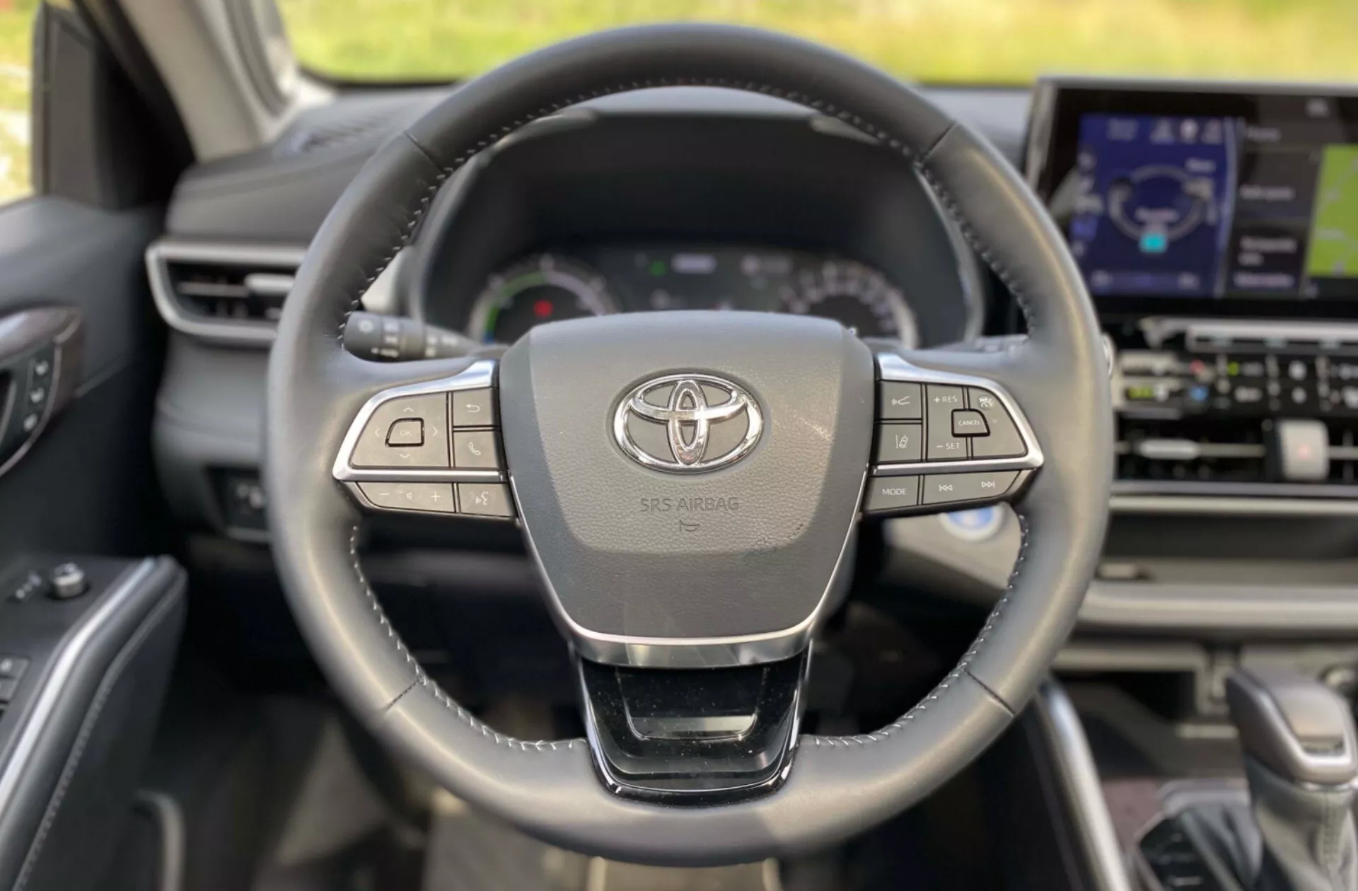 Interiör av en 2021 Toyota Highlander Business Edition från förarplatsens perspektiv, med en modern, läderklädd treekrad ratt med flera kontrollknappar, Toyota-logotypen och SRS AIRBAG-inskriften. I bakgrunden syns instrumentbrädan med en digital displaykluster och större infotainmentskärm.