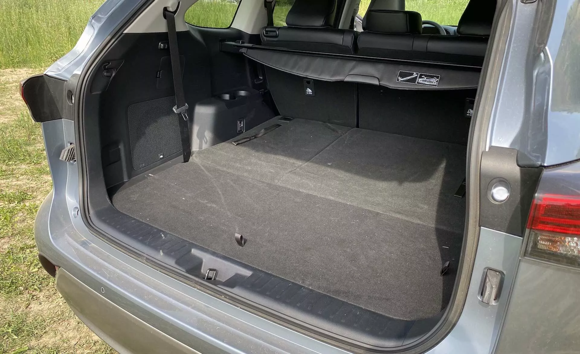 Närbild av det öppna lastutrymmet i 2021 års Toyota Highlander 2.5 Hybrid Business Edition med vertikal baklucka och rymligt bagageutrymme. Bildvinkeln från passagerarsidan visar en ren, mattbeklädd yta, med eventuellt nedfällda baksäten för att öka lastutrymmet. Synliga funktioner inkluderar en innerbelysning, ett 12V-uttag, en dragflik för ytterligare förvaring, fästen för barnstolar och en del av det moderna LED-bakljussystemet.