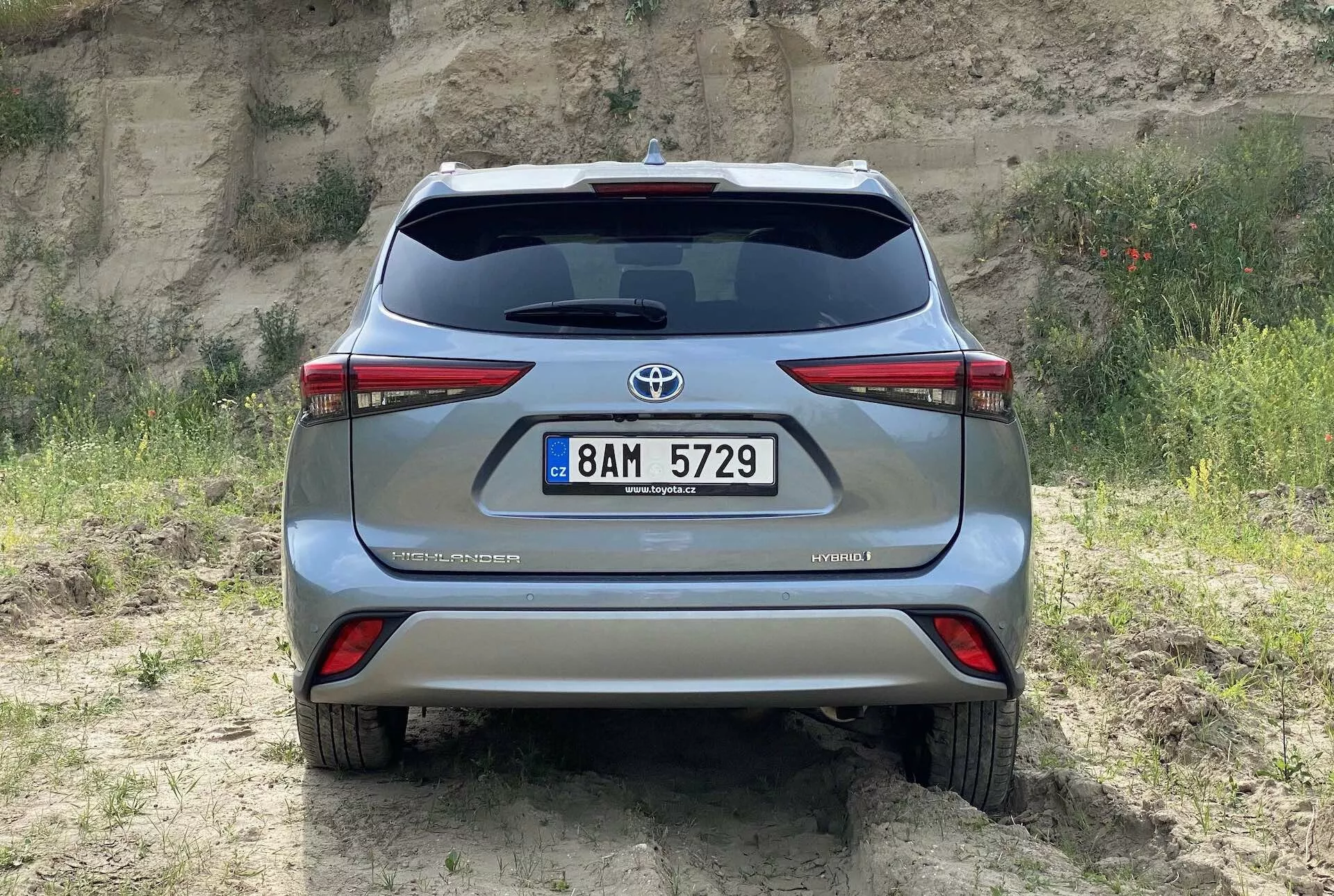 Bakifrån vy av en silverfärgad 2021 Toyota Highlander 2.5 Hybrid Business Edition fångad i ögonhöjd, som visar bakluckan med Toyota-emblemet, bakljus, dubbelt avgasrör, registreringsskylt i mitten av den bakre stötfångaren och hybridmärket – allt placerat på ett naturligt terräng med gräs och jord, som framhäver dess terrängkapacitet.