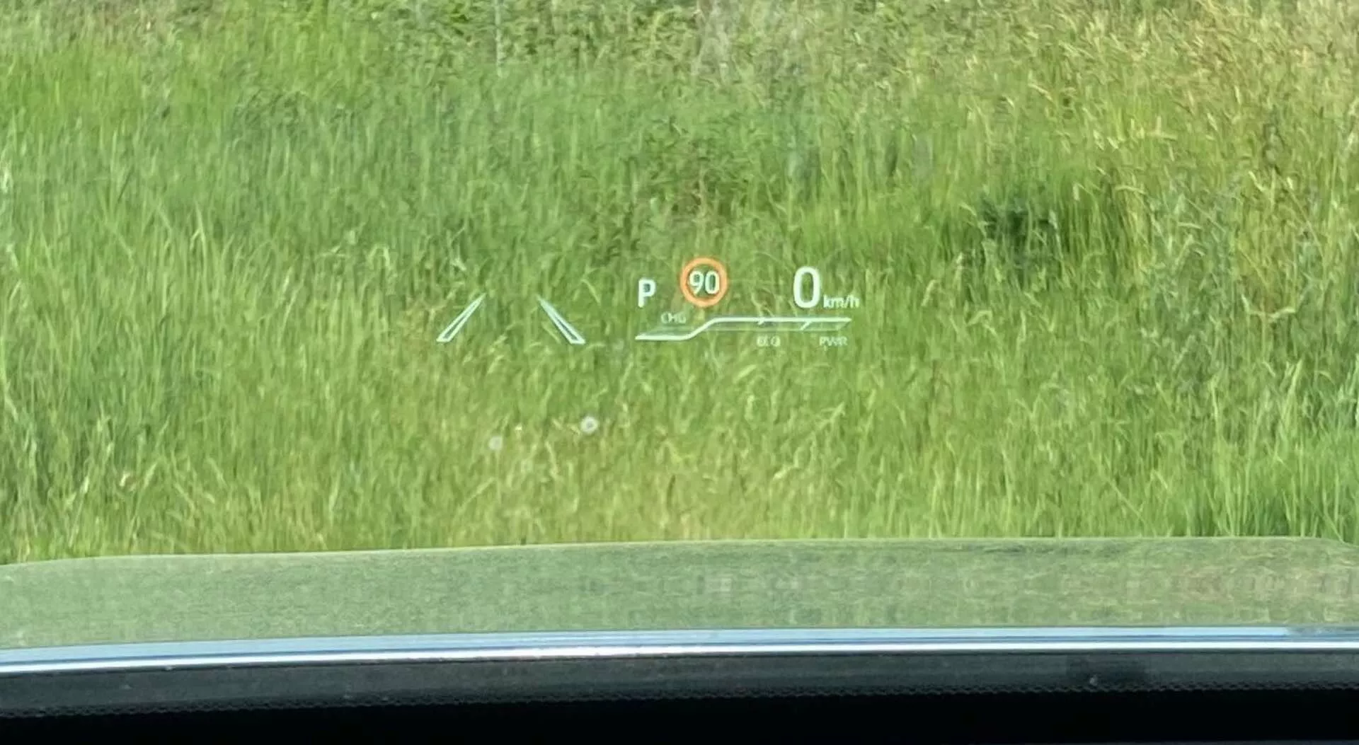 Interiör vy av en Toyota Highlander 2021 2.5 Hybrid Business Edition med fokus på head-up display (HUD) projicerat på framrutan. HUDen visar viktig körinformation: en 'P' för 'Park' växelindikator, en hastighetsgränsskylt med '90', en aktuell hastighet på '0 km/h' som indikerar att bilen står stilla och symboler förmodligen relaterade till parkeringsassistanssystem eller körfältsassisteringsindikator. Scenen är tagen något till höger om förarsätet och visar utsikt mot ett gräsområde utanför.
