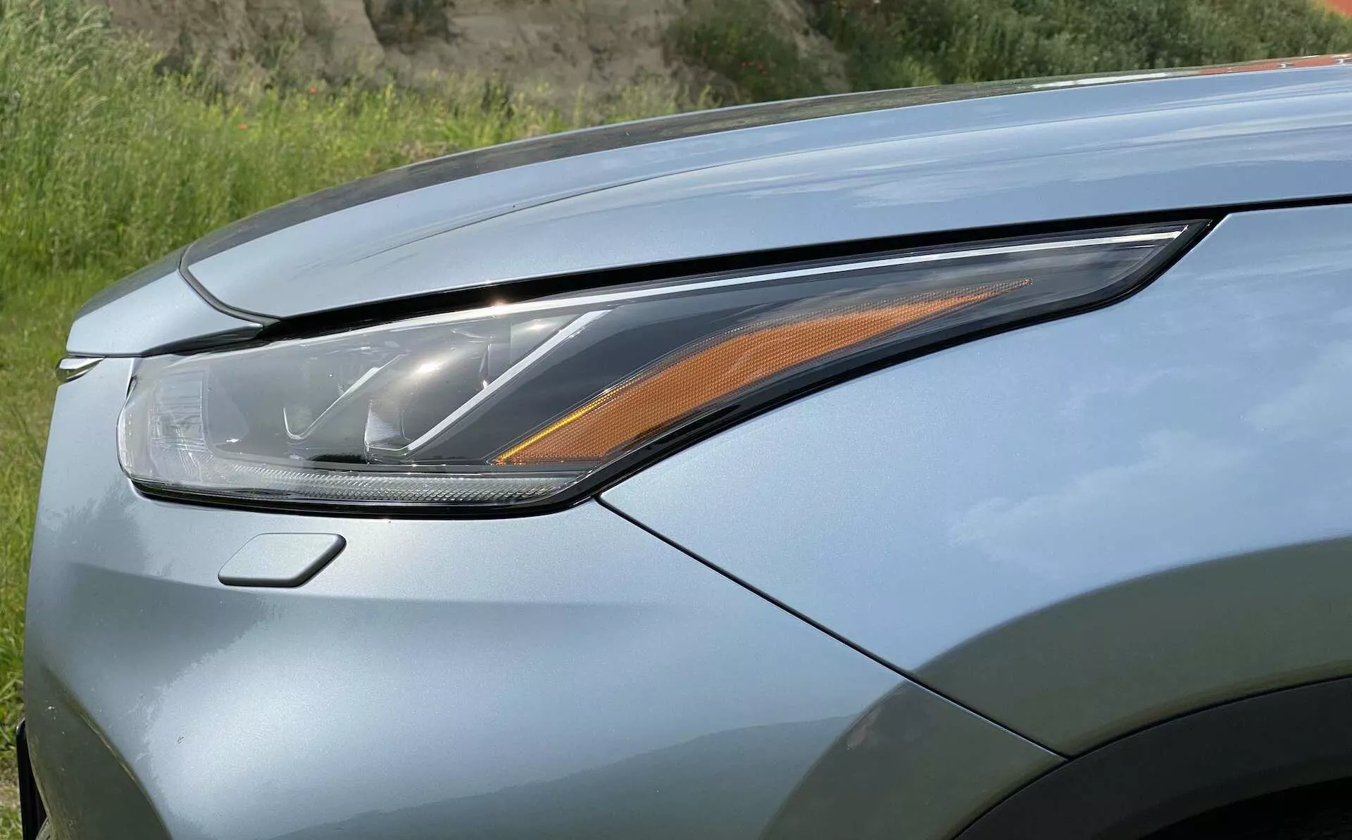 Närbild av den vänstra främre delen av en Toyota Highlander 2.5 Hybrid Business Edition 2021, med fokus på strålkastaren och en del av motorhuven. Bilden är tagen från en låg, lätt uppåtvinklad och framåtriktad vinkel, vilket framhäver den moderna LED- eller laserstrålkastaren med flera ljuselement, inklusive en amberblinkers. Motorhuven har en metallisk lack med möjlig pärlemofinish och aerodynamiska linjer, vilket betonar den avancerade belysningsteknologin och den lyxiga designen.