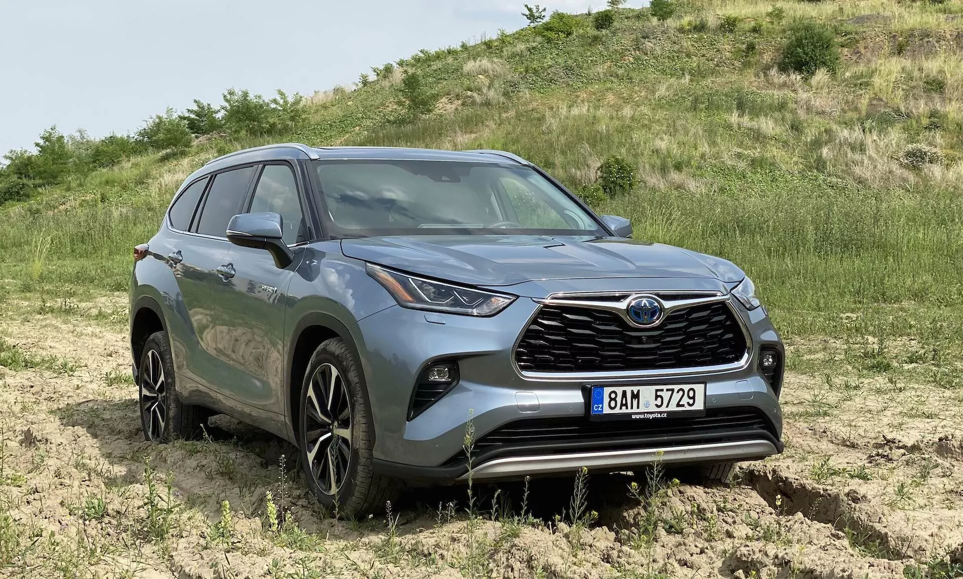 En tre-kvart framåtvänd vy av en grå Toyota Highlander 2.5 Hybrid Business Edition SUV placerad utomhus i ett off-road-område med gräs och jord. Bilden lyfter fram den framträdande frontgrillen med varumärkesemblemet, de distinkta LED-strålkastarna som går runt till sidan, fler-eker legeringshjul på cirka 18-20 tum, skulpterade karosslinjer med konturerade skärmar, den förhöjda höjden som antyder fyrhjulsdrift, dimljus på främre stötfångaren, sidobackspeglar med integrerade blinkers och rökfärgade glas på de bakre fönstren. Bilden ger en tydlig vy av dessa funktioner på ögonhöjd med naturlig belysning som visar bilens färg och designdetaljer.
