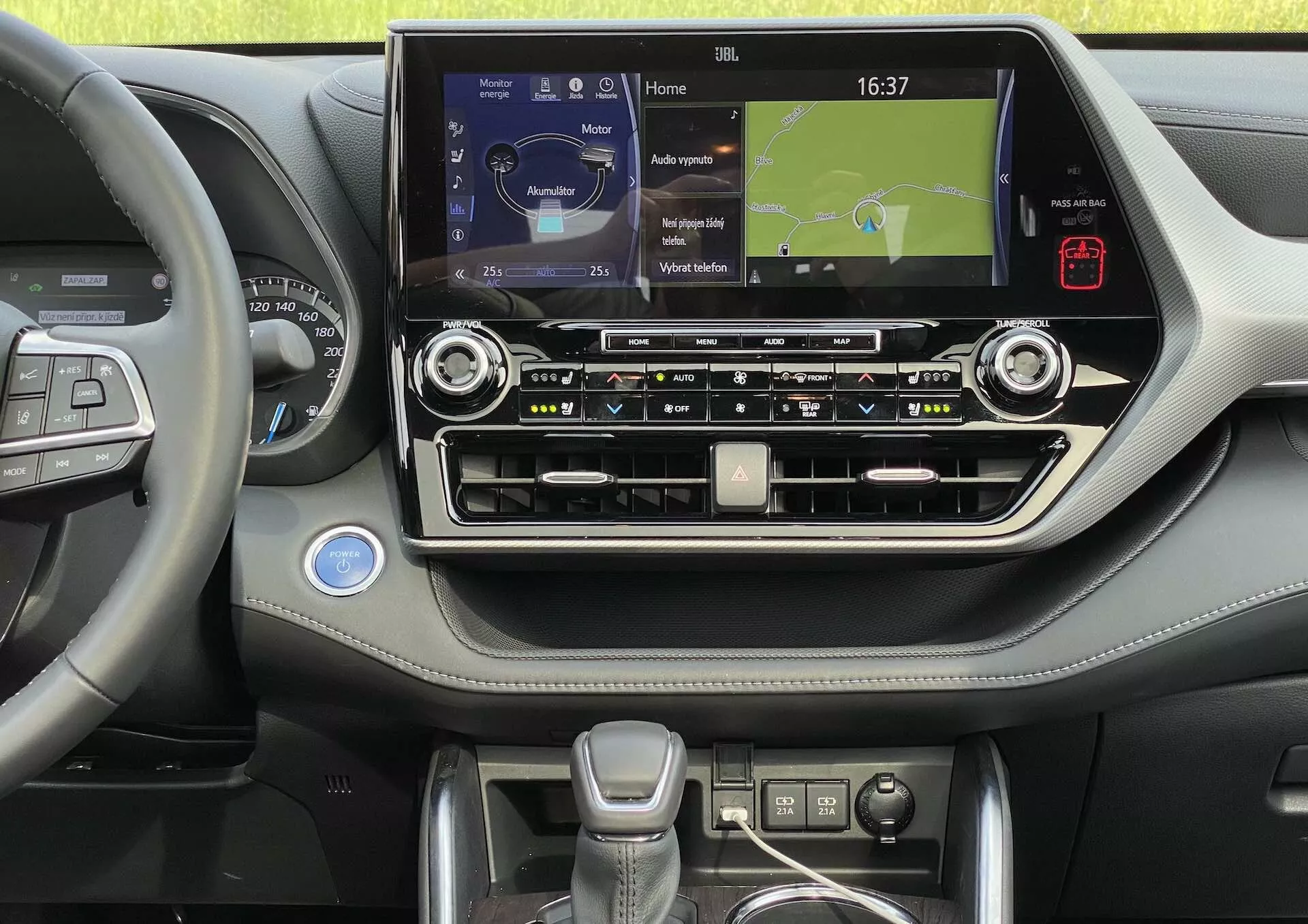 Interiören i en Toyota Highlander 2021, med fokus på mittkonsolen och förarsidan. Bilden, tagen från ögonhöjd, visar ratten med multifunktionsknappar, som antyder funktioner som farthållare, ljudsystemkontroll och handsfree-samtalshantering. Mittkonsolen domineras av en stor pekskärm som visar en navigeringskarta, vilket indikerar media- och telefonanslutningar. Under pekskärmen finns knappar och rattar för klimat- och ljudsystem, tillsammans med en analog klocka, metalliska accenter och en läder- eller mjukdaskinläggning. Växelspaken är modern och ergonomisk, med ytterligare kontroller runt omkring, sannolikt för körlägen. Längst ner i bilden finns antydan om mugghållare eller förvaringsutrymme. Interiören är ren, välbelyst och väl underhållen.