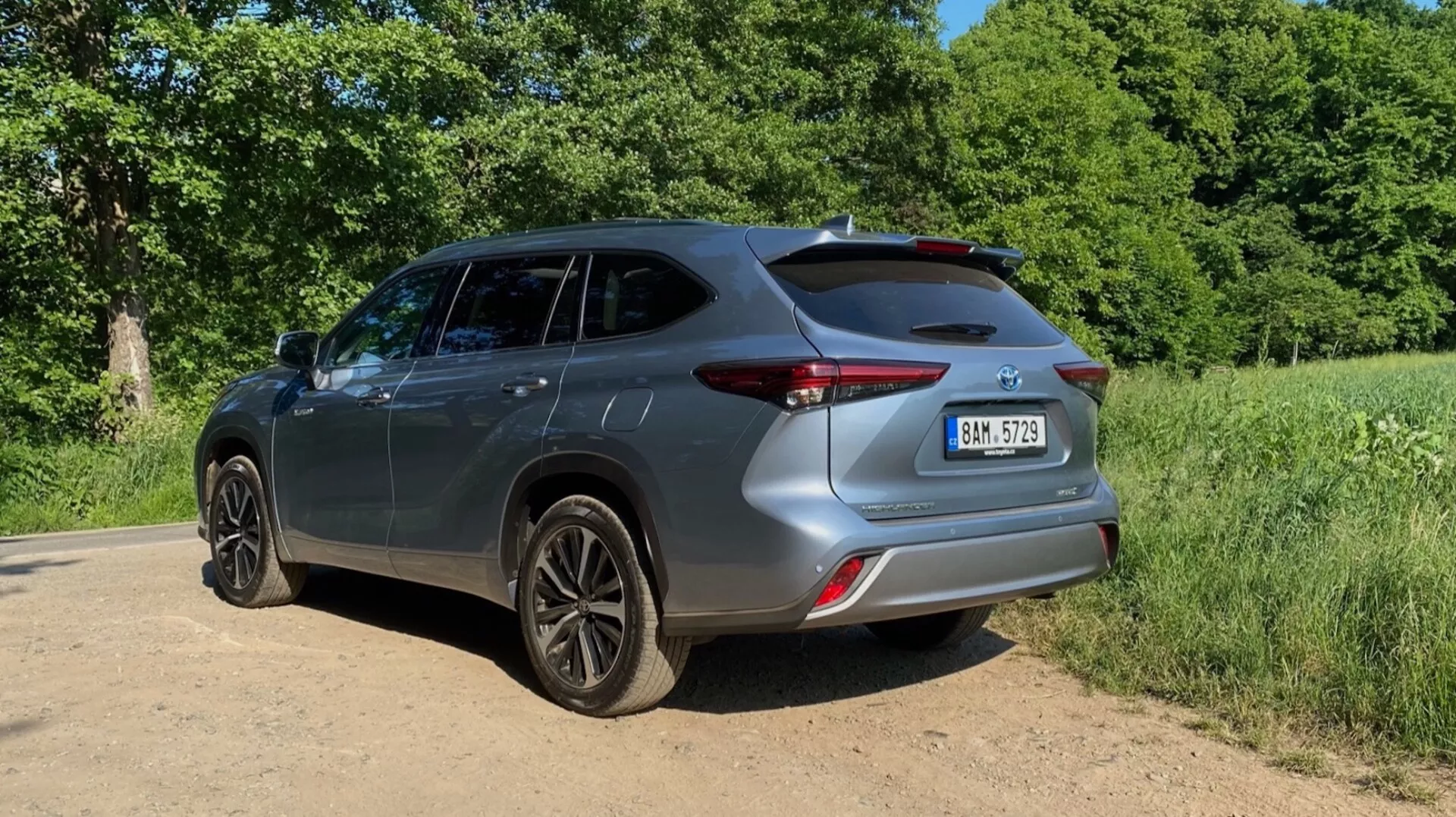 Toyota Highlander 2021 Hybrid Business Edition visad från en bakre trekvartsvy, som framhäver bilens högra sida och bakände. Marknivåbilden betonar de mörkfärgade alluminiumfälgarna och den totala höjden på bilen. Den bakre stilen, med eleganta bakljus, konturerad stötfångare och baklucka med en bakre spoiler, är framträdande. Bilden visar också utformningen av dörrarna, sidorutorna och speglarna. Fordonet är placerat på en grus- eller grusstig med grönska och träd i bakgrunden. Det soliga ljuset kastar skuggor framåt, vilket tyder på att solen är bakom kameran.