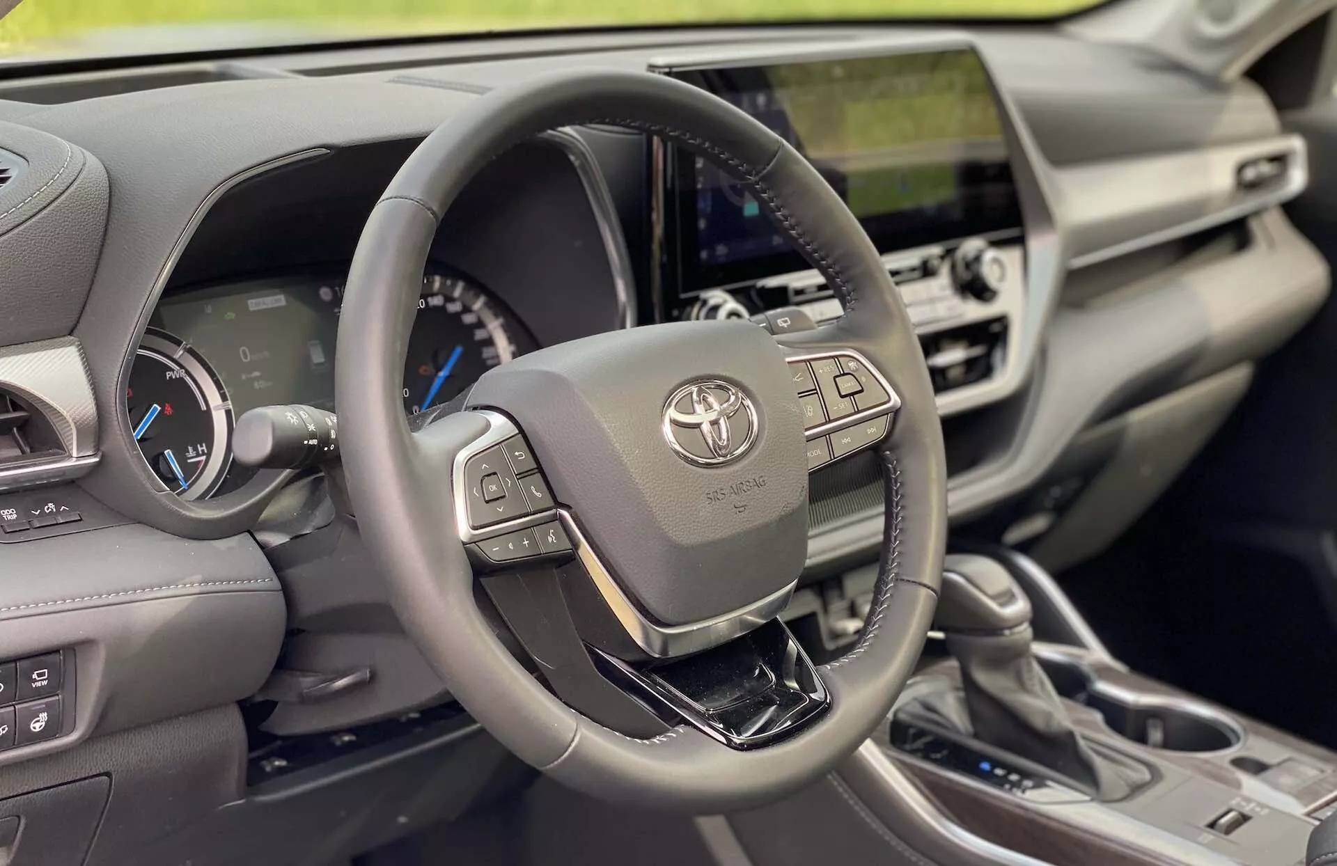 Interiörvy av en 2021 Toyota Highlander 2.5 Hybrid Business Edition, med fokus på förarsidan. Bilden, tagen från en aningen ovanför och från fram-höger sida, visar tydligt Toyota-emblemet, multimedia- och informationsdisplaysknappar till vänster och knappar för den adaptiva farthållaren och handsfreekommunikation till höger. Instrumentpanelen bakom ratten inkluderar en digital skärm med blått tema och två analoga mätare, vilket indikerar ett hybridfordon. Centralpanelen inkluderar en integrerad pekskärms-infotainmentenhet, luftventiler och klimatanläggningskontroller. Materialen runt cockpiten antyder en modern design med betoning på funktionalitet och en touch av lyx, med läder- eller konstläderklädsel, en metallisk växelspak för automatlådan och olika körkontroller. Naturligt ljus belyser interiören, framhäver de intrikata detaljerna och texturerna kring ratten och instrumentpanelen.