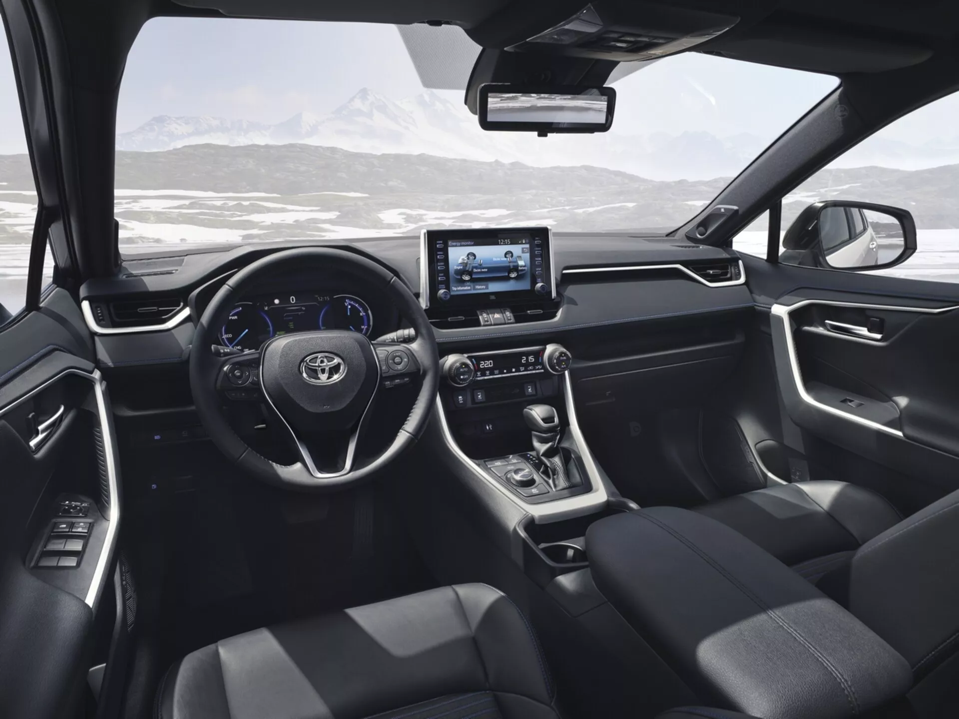 Interiörvy av Toyota RAV4 2019 från baksätet, visar den moderna designen och tekniken inklusive ratten med kontrollknappar, digital display, pekskärmsinfotainmentsystem, växelspak för automatisk växellåda, mörkt klädda säten och dörrpanelkontroller med en scenisk snöig bergslandskap synlig genom vindrutan.