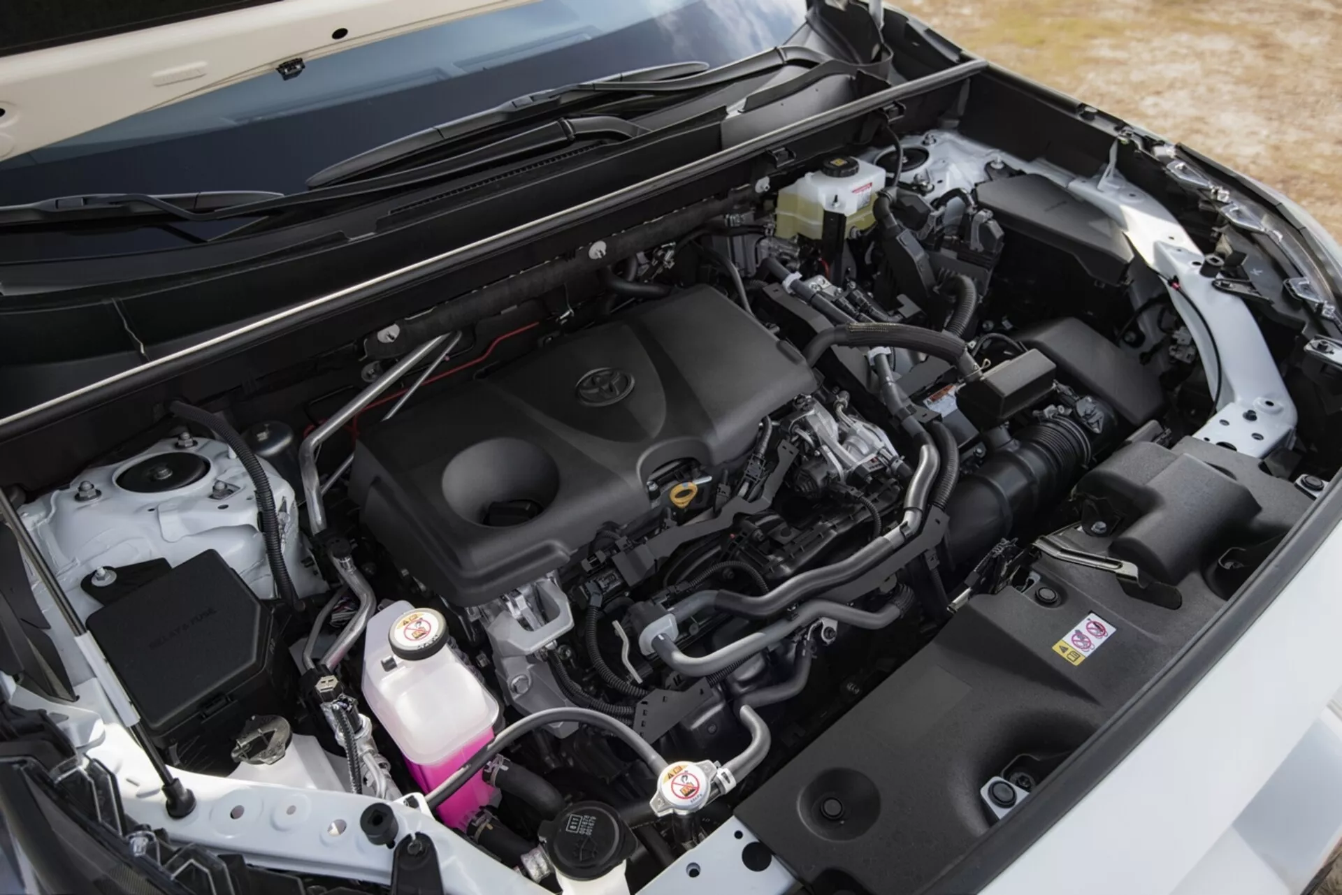 Närbild på motorutrymmet i en Toyota RAV4 från 2019, som framhäver den rena, moderna designen och komponenter såsom motorlocket med märkesemblem, luftintagssystemet, batteriet, bromsvätskebehållaren och kylvätskebehållaren, tillsammans med olika rör och kabelhärvor, under det öppna motorhuven från en sned vinkel.