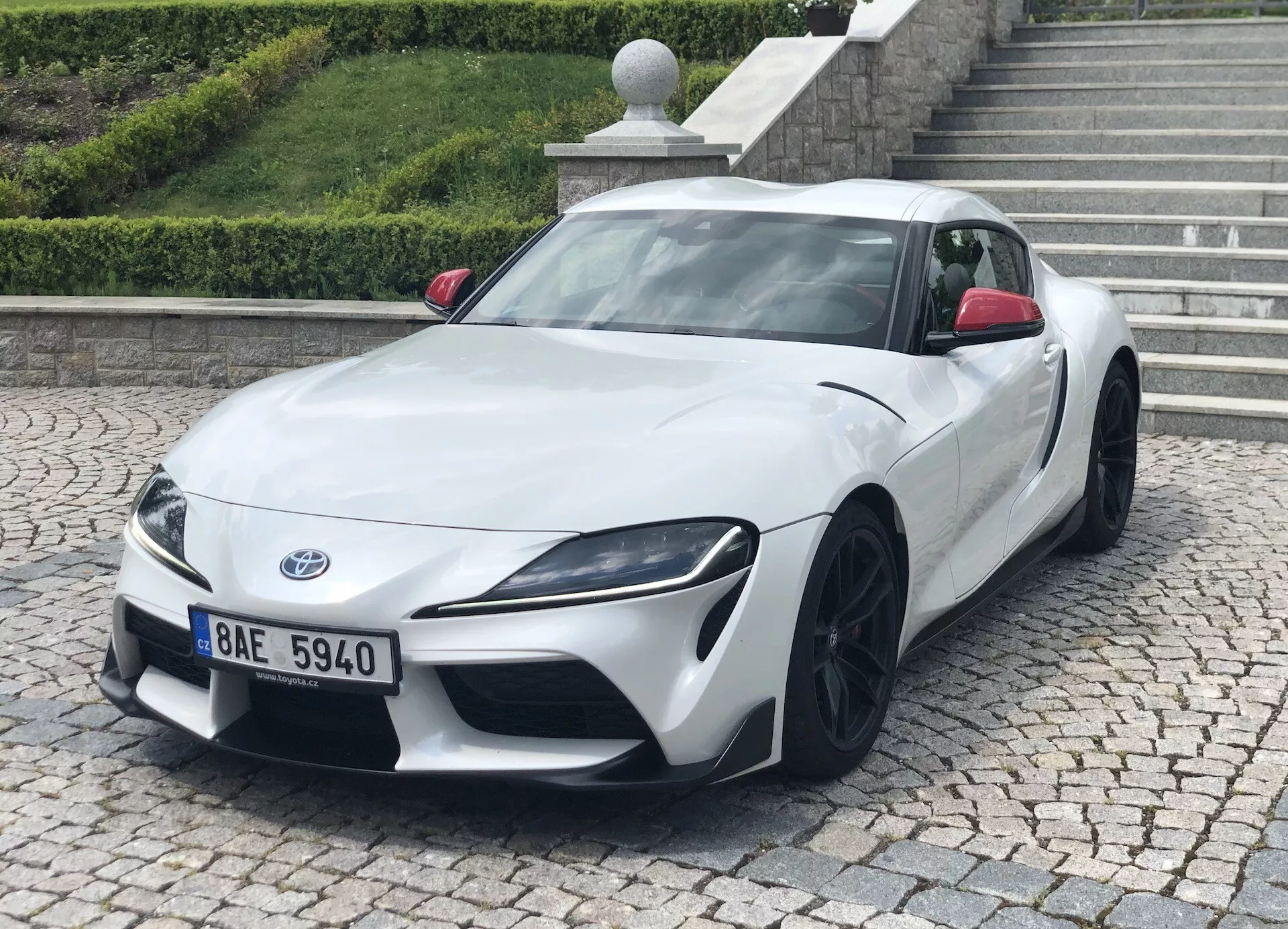 Bilden visar en vit Toyota Supra 2019 (toyota-gr-supra-2-0-pure-automatic) sedd från en trekvartsvinkel framifrån. Bilens låga och breda hållning, skarpa vinklade strålkastare och svarta flerspokeslättfälgar är tydligt synliga. Fotot framhäver den eleganta designen, aerodynamiska konturer och prestandaestetiken. Sportbilen är parkerad på trottoaren med en kantsten och en trappa i bakgrunden, vilket tyder på en stads- eller bostadsmiljö.