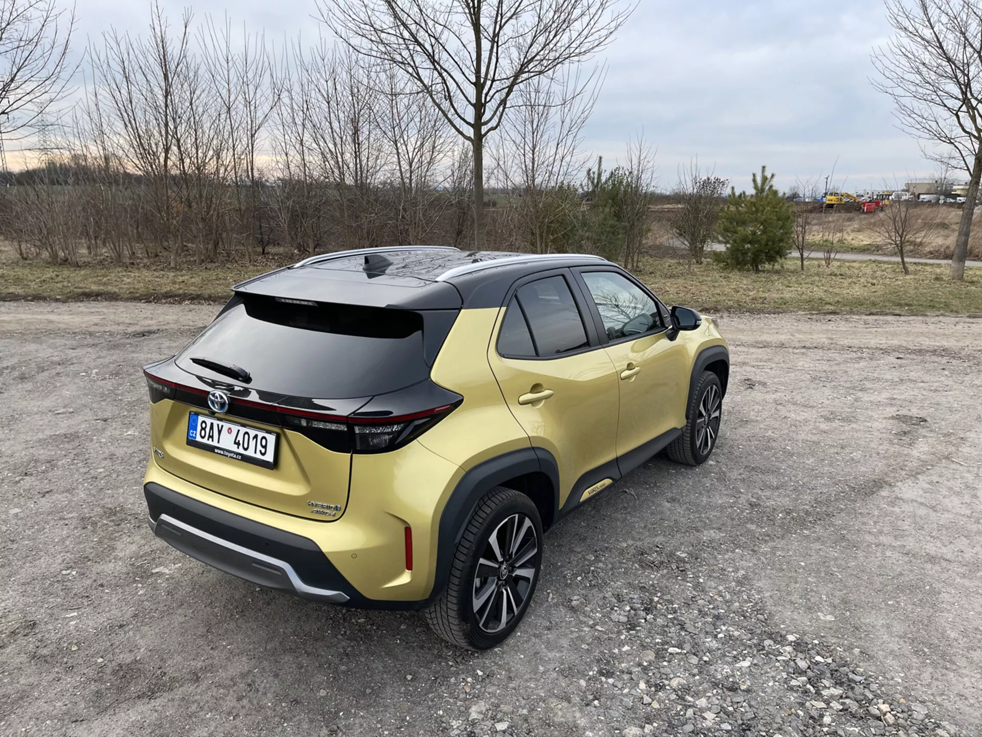 Toyota Yaris Cross 2021 fångad från en bakre trekvarts vy som framhäver dess aerodynamiska spoiler, metalliskt guld färg, och LED-bakljus, parkerad på en opackad yta med naturliga och industriella bakgrunder.