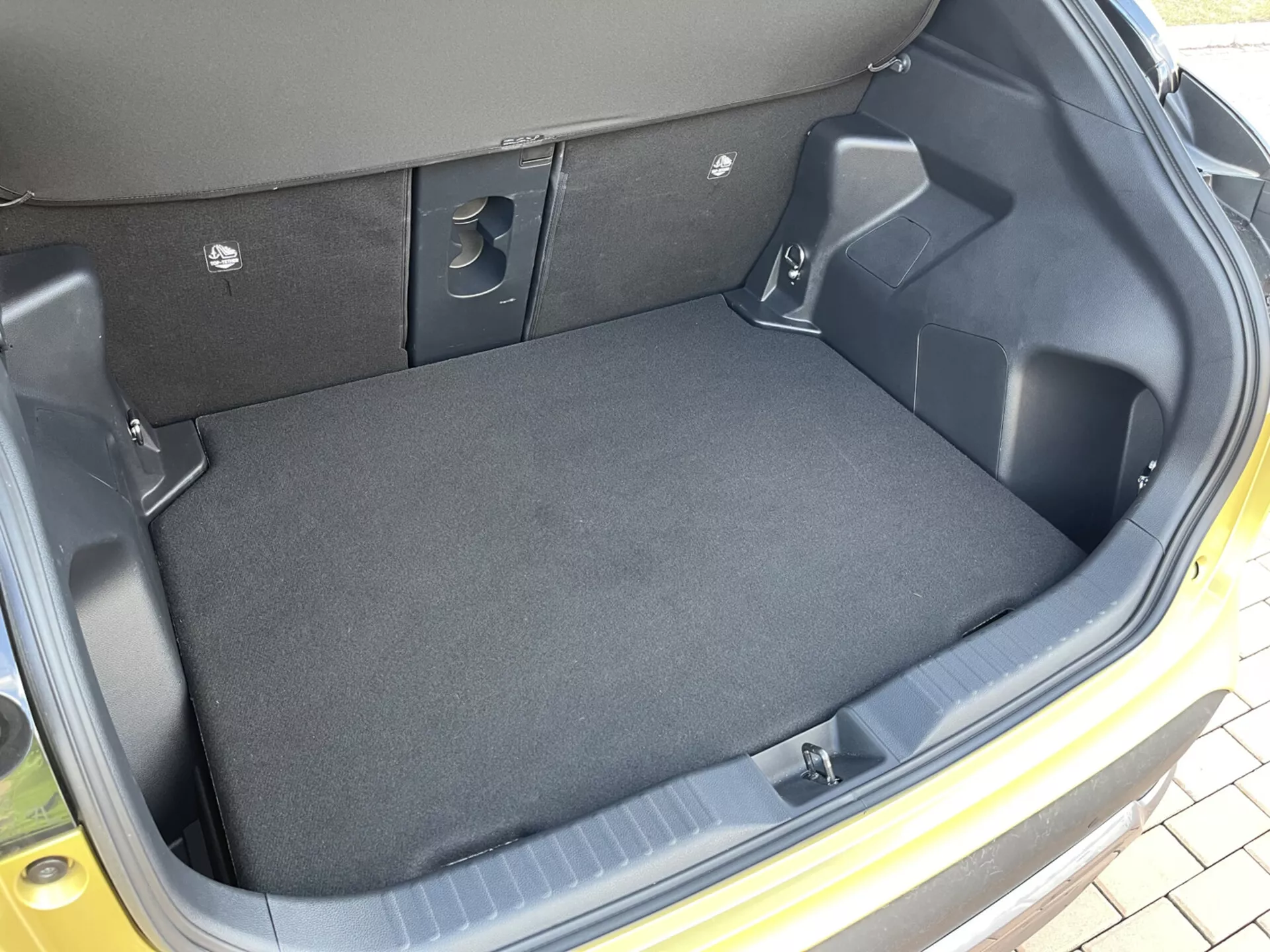 Interiörvy av bagageutrymmet i Toyota Yaris Cross 2021 som visar ett tomt, mattbelagt lastutrymme med formgjutna plastsidor, fördjupningar för förvaring, utskärningar för tillgång till utrustning och indikationer på fällbara baksäten