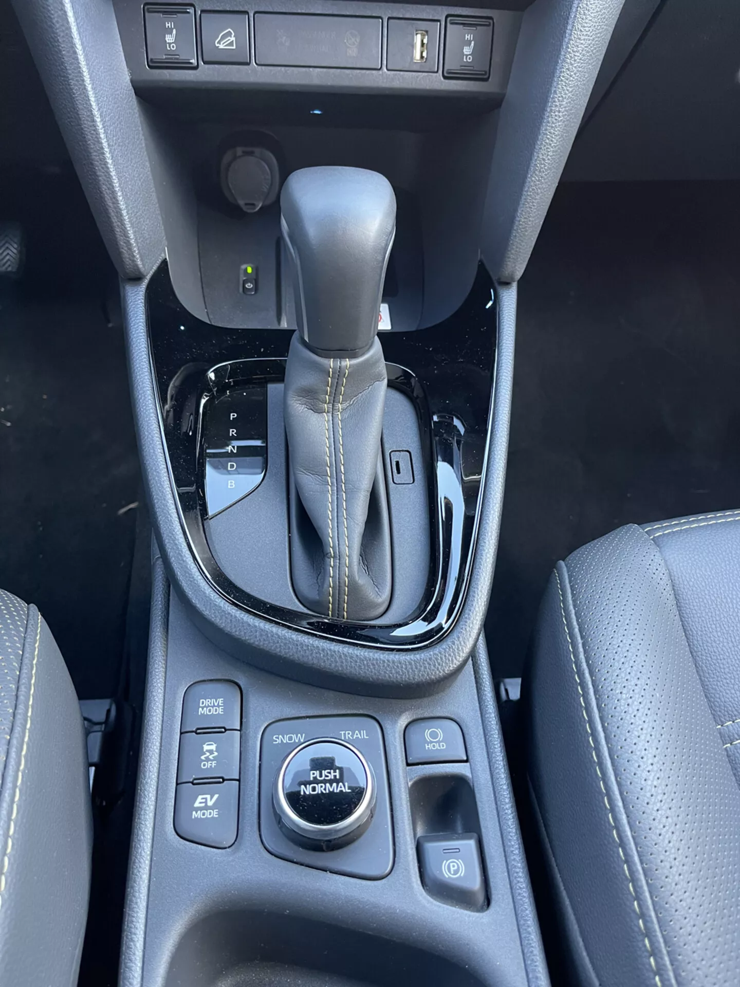 Interiör av Toyota Yaris-Cross 2021 som visar mittkonsolen, automatisk växelspak i parkläge med metallinlägg och blanksvart infattning, valknappar för körningsläge inklusive SPORT, ECO, EV, NORMAL, slirskyddskontroll, sätesvärmningskontroller och ett 12V-uttag, vilket betonar designkvalitet och avancerade fordonstekniska funktioner.