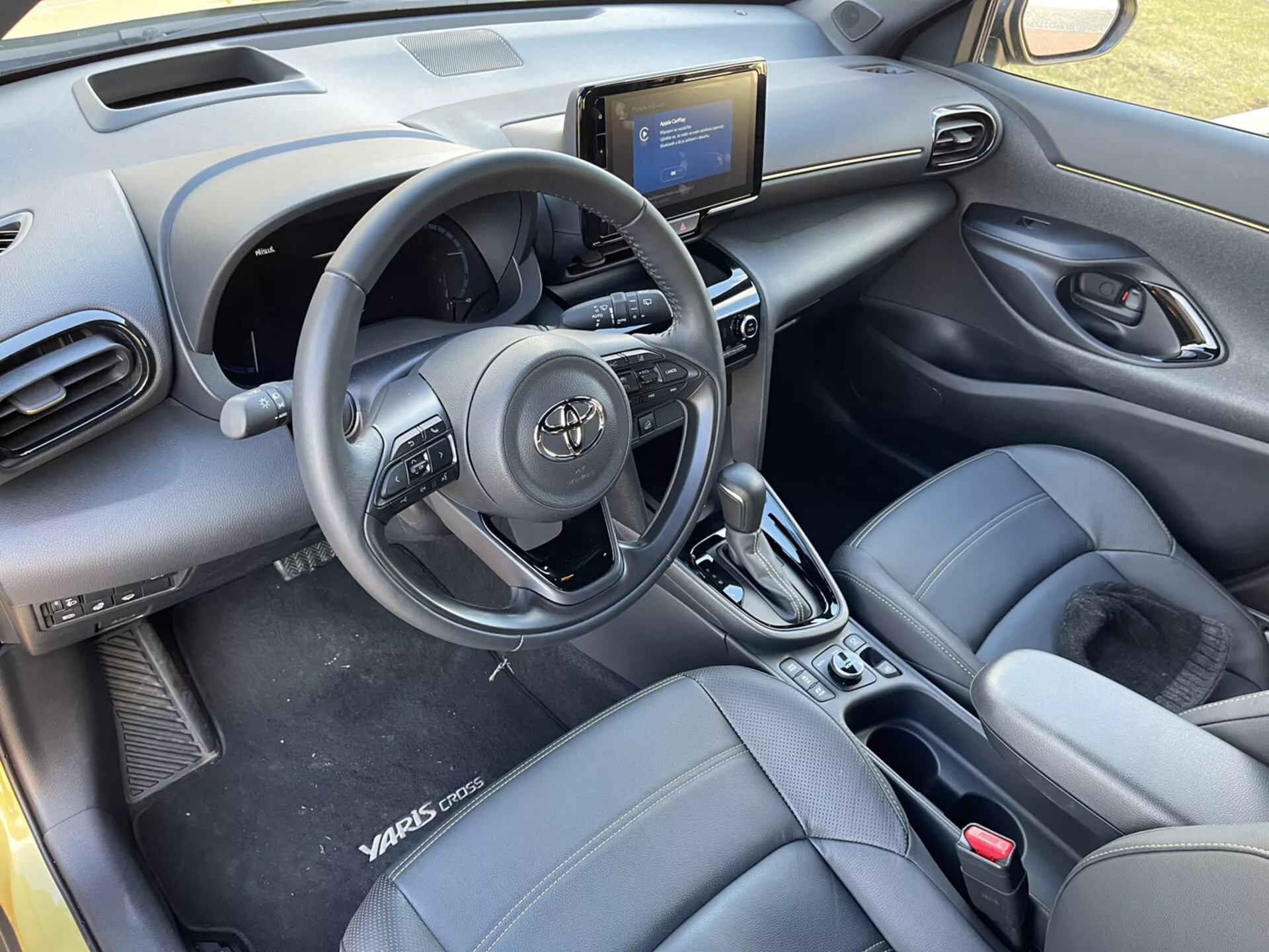 Interiörvy av Toyota Yaris-Cross 2021 från baksätena, framhäver ratten med kontroller, luftventiler på instrumentbrädan, mittkonsol med infotainmentsystem, luftkonditioneringskontroller, automatisk växelspak, läderliknande främre hinkstolar med Yaris Cross-märkning, dörrpaneler med elkontroller, och en modern, elegant kupédesign.