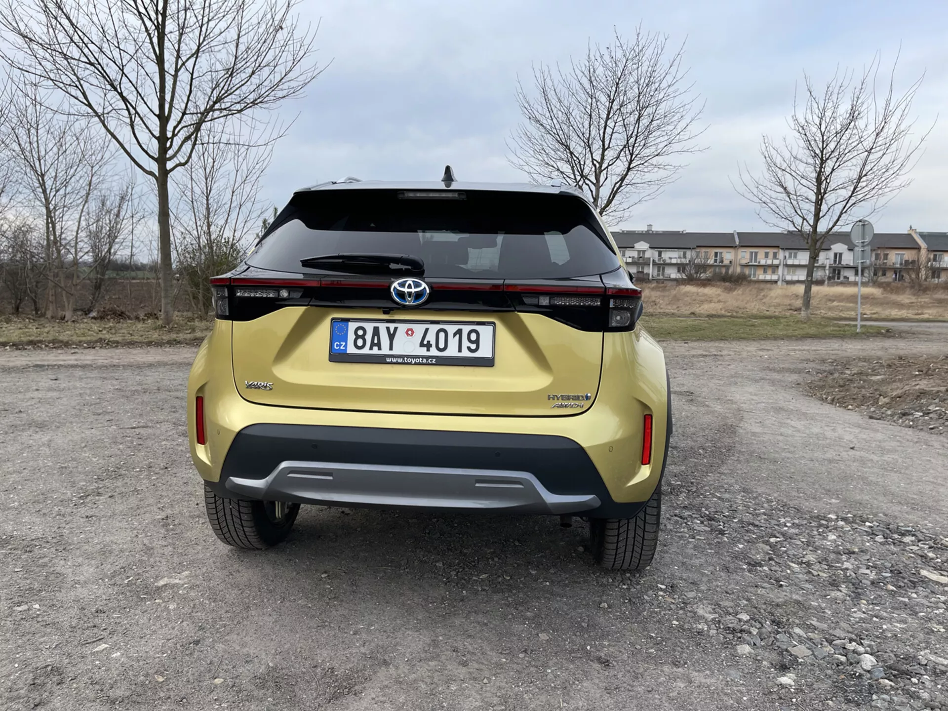 Baksida av en gul Toyota Yaris Cross 2021 tagen i ögonhöjd som visar dess moderna design, horisontella bakljusenhet, Toyota-emblem, modellnamn Urban Cruiser, registreringsskylt, bakre stötfångare med kontrasterande design med integrerade parkeringssensorer, och bagagehandtag mot en förorts bakgrund.