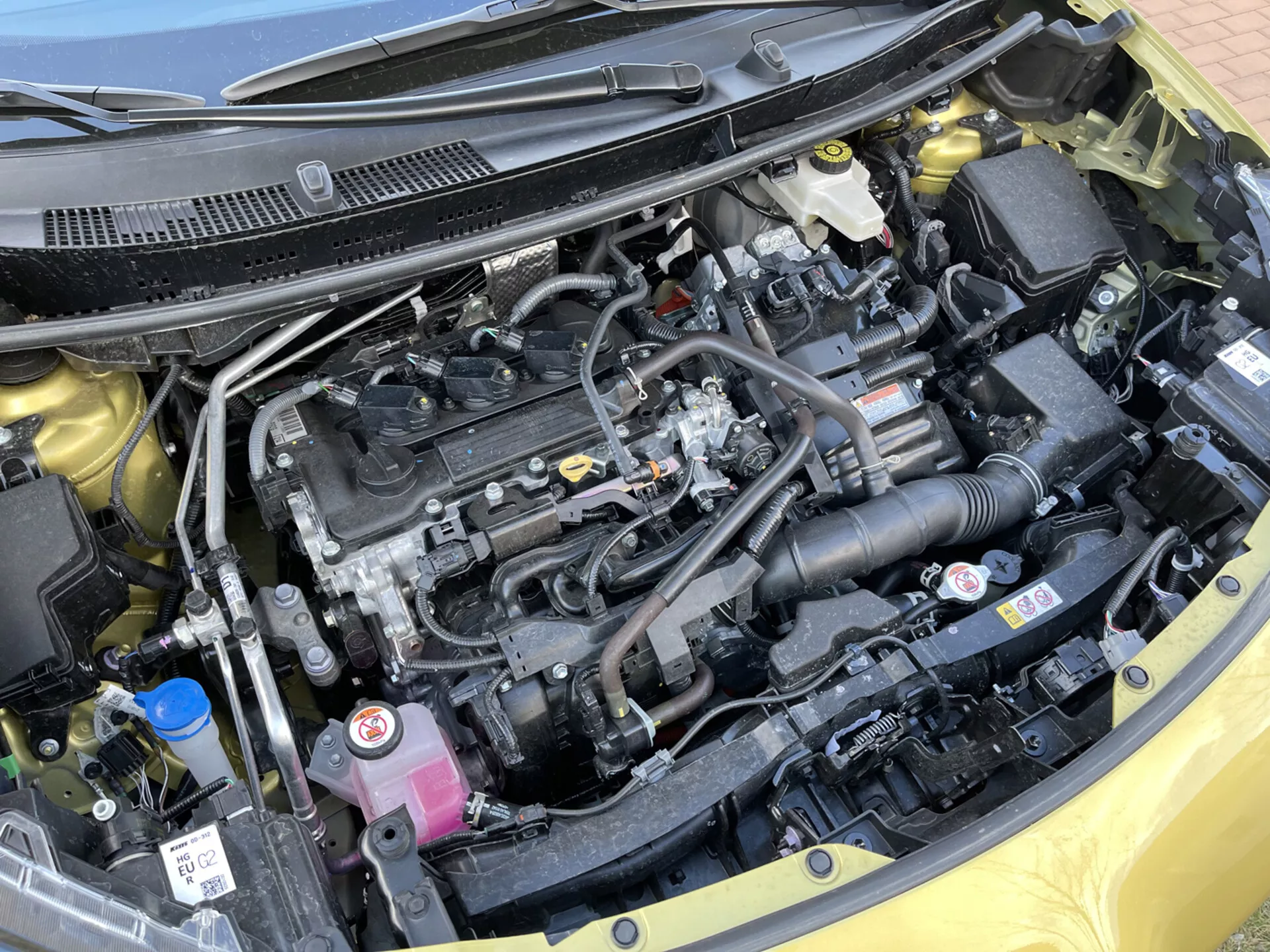 Närbild av motorutrymmet i Toyota Yaris-Cross 2021 som visar en rak motorblock, svart plastmotorlock, luftintagssystem, kylarvätske- och bromsvätskebehållare, elektriska komponenter, batteri, kylare och en snyggt organiserad layout med säkerhetsetiketter.