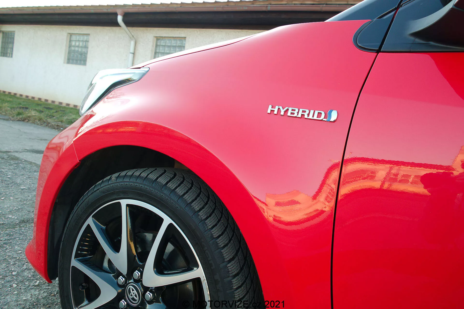 Närbild av en röd Toyota Yaris 2020, med fokus på framflygplanet och den flerspakiga lättmetallfälgen. Hybridmärket är synligt och framhäver fordonets miljövänliga teknik. Den glansiga lacken reflekterar solljus, vilket betonar bilens högkvalitativa finish och moderna, aerodynamiska design.