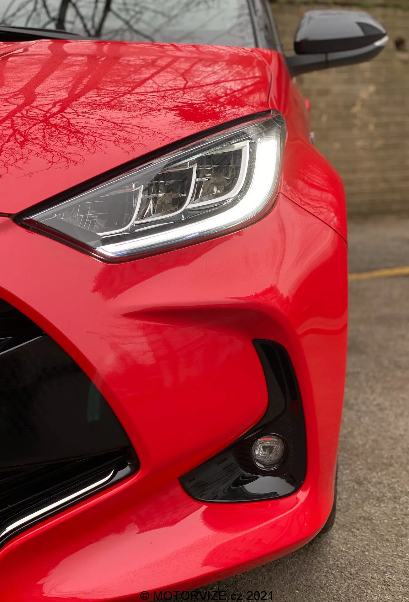 Närbild av Toyota Yaris 2020, som visar framsidan på förarsidan från en låg vinkel. Bilden framhäver bilens ljusa, glansiga röda färg, slanka LED- eller HID-strålkastare med distinkta dagsljuskörlampor och delvis synlig sidospegel i samma färg som karossen med integrerad blinkers. Den aerodynamiska designen betonas genom de skulpterade linjerna och kurvorna runt strålkastaren och motorhuven. En rund dimljus eller sensor på främre stötfångaren tyder på avancerade säkerhetsfunktioner. Avsaknaden av rörelseoskärpa indikerar att bilen är stillastående, och en vattenstämpel 'MOTORZETZ. 2021' syns längst ned till höger.