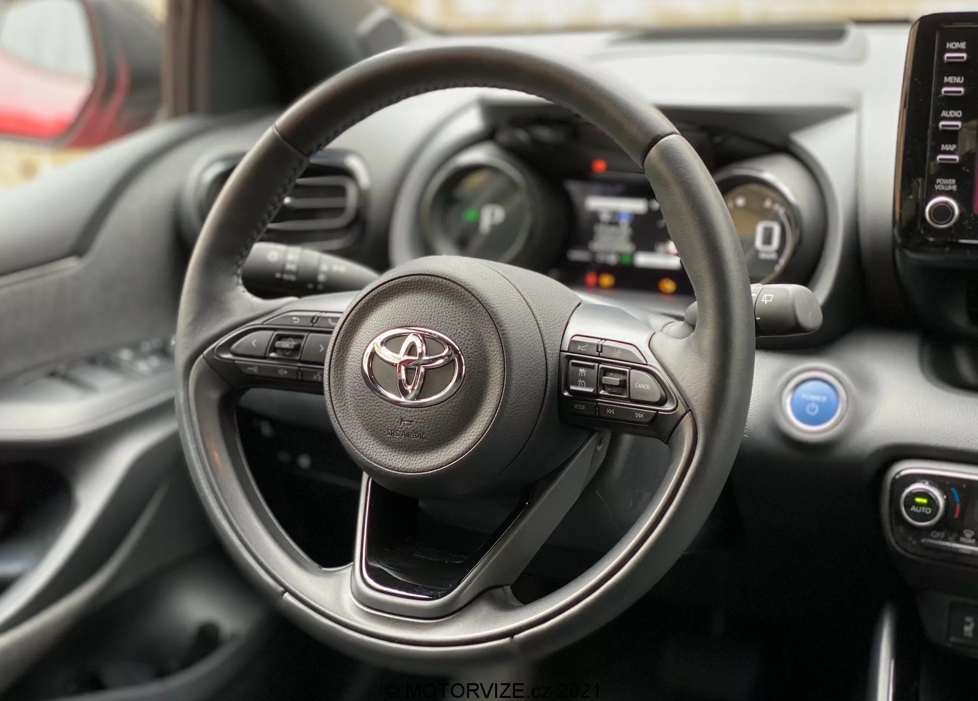 Interiörvy av en Toyota Yaris 2020 med fokus på ratten och instrumentpanelen från förarsätets perspektiv. Ratten har Toyotas logotyp och synliga kontroller för infotainmentsystemet och förarassistanssystem. Bakom ratten syns delar av den digitala instrumentpanelen och mittkonsolen med klimatkontrollreglage och luftventiler.