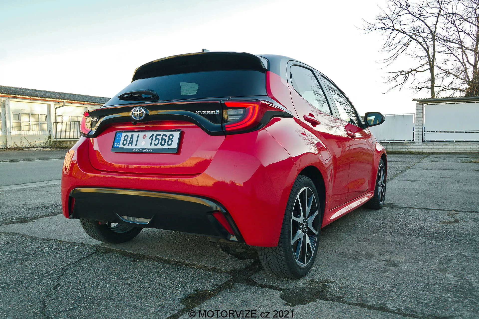 Livlig röd Toyota Yaris 2020 personbil fotograferad från en bakre trekvartsvinkel, som framhäver LED-bakljus, bakre spoiler, Hybridmodellemblem, fler-ekrade lättmetallfälgar och synliga förarassistansfunktioner som parkeringssensorer och backkamera, fångat ur ett något lägre bilhöjdsperspektiv.