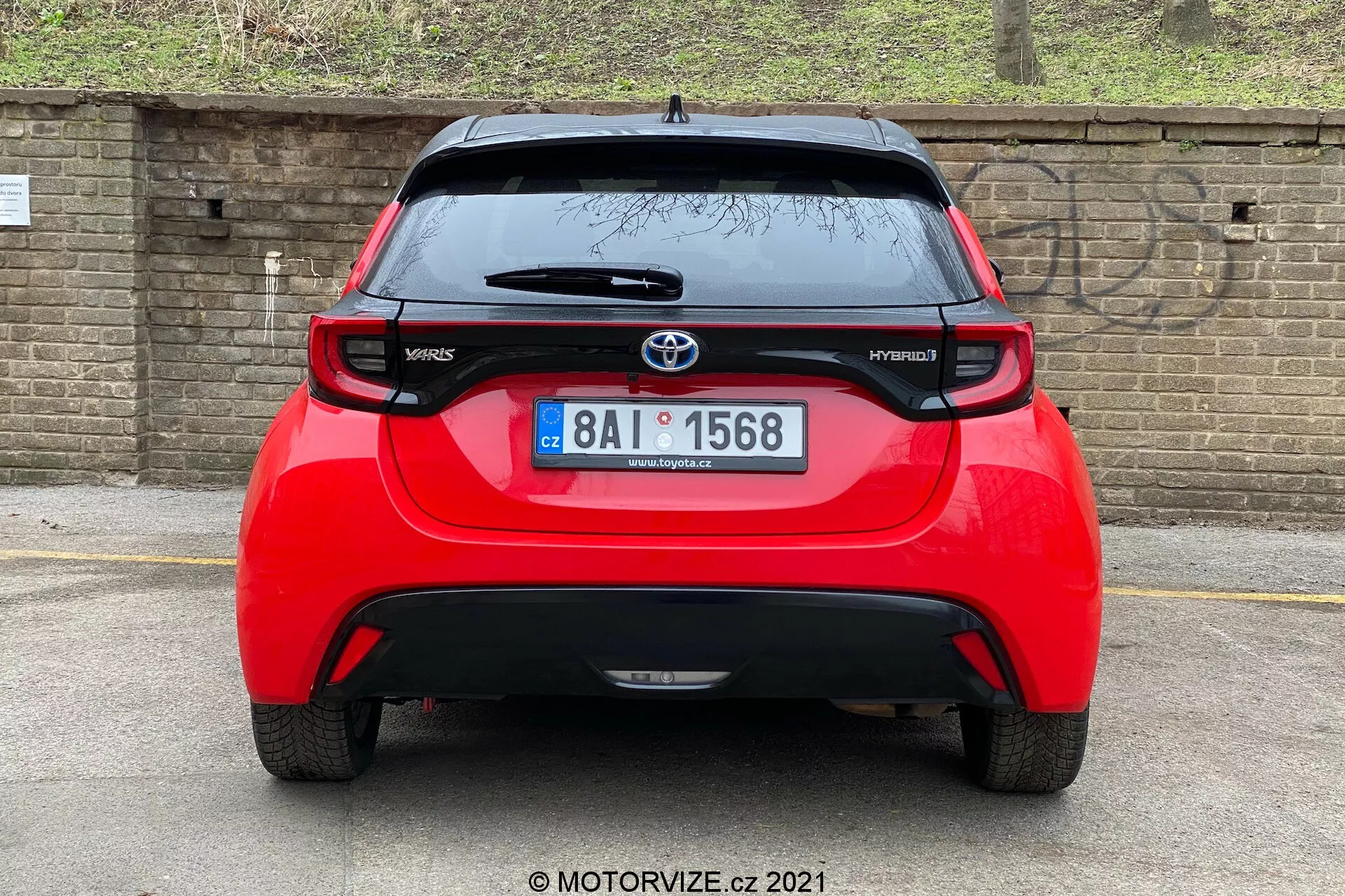 Bakre vy av en röd Toyota Yaris Hybrid från 2020, centrerad och tagen ur en direkt vinkel. Bilden visar hela fordonets bredd och framhäver Toyota-emblemet, modellnamnen 'Yaris' och 'Hybrid', en spoiler, takmonterad antenn, en högmonterad bromsljusremsa och distinkta bakljus. Bakrutan med en vindrutetorkare och den nedre stötfångardelen, som eventuellt innehåller sensorer eller reflexer, är också synliga. I bakgrunden finns en tegelvägg eller ett staket och lite växtlighet, vilket tyder på en lägre parkerad bil eller en lägre kameravinkel.