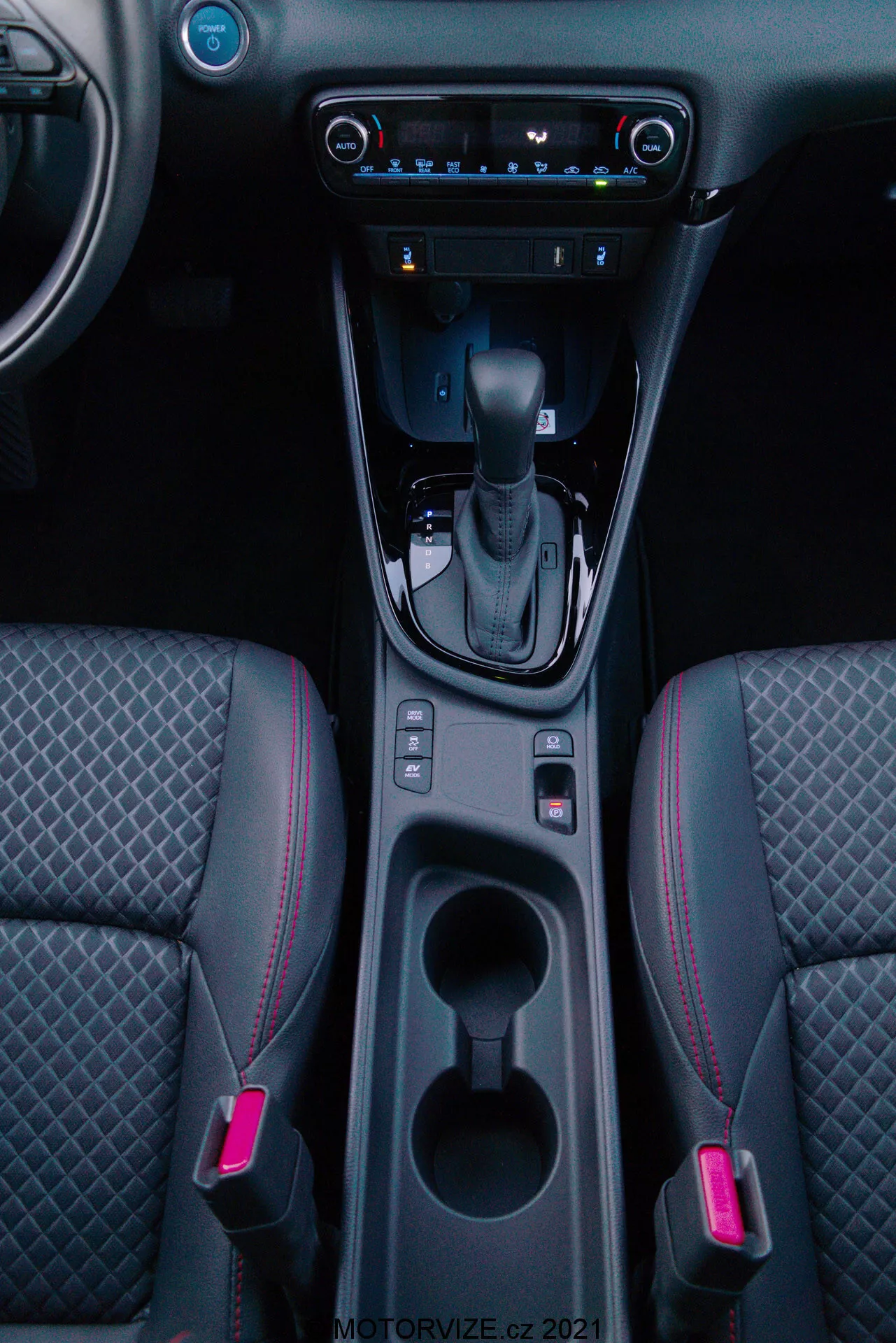 Vertikal interiörbild av en 2020 Toyota Yaris med fokus på mittkonsolen, med växelspaken i 'P'-läge, elektrisk parkeringsbromsknapp, två upplysta mugghållare, förvaringsutrymme, digital klimatkontroll, pekskärmsmultimediasystem, USB-port, luftventiler och tvåfärgade framsäten med diamantmönstrad sömnad och röda accentsömmar.
