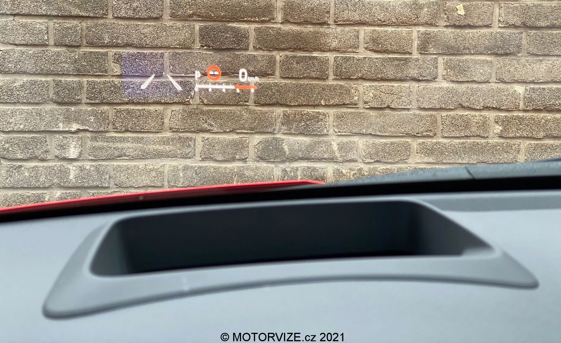 Närbild från insidan av en 2020 Toyota Yaris som visar en delvis vy från förarsätet mot bakre delen. Fokuset är på den bakre hyllan med ett högmonterat bromsljus och integrerad bakspoiler. Bilden visar även en del av innermaterialet, medan en tegelvägg utanför antyder att bilen är parkerad nära en byggnad.