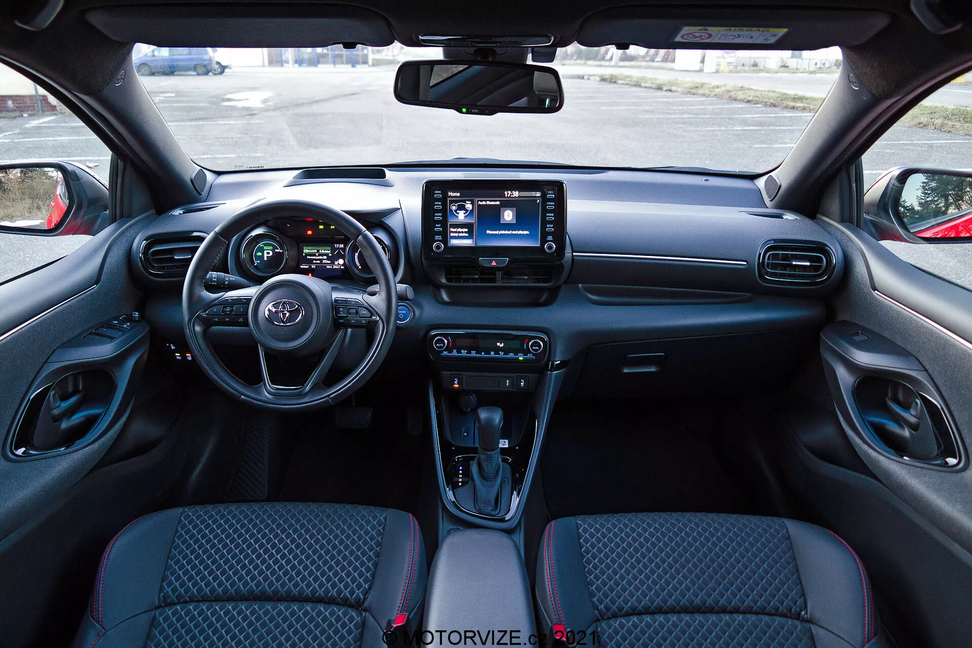 Interiörvy av en Toyota Yaris 2020 från förarens perspektiv, med en modern instrumentbräda som har en ratt med inbyggda kontroller, en delvis synlig instrumentpanel med digitala skärmar och ett centralt infotainmentsystem med pekskärmsgränssnitt. Instrumentpanelen inkluderar ventilationsöppningar och klimatkontrollknappar, medan mittkonsolen har kontroller för körlägen, en automatisk växelspak med läderliknande skydd, mugghållare och ett förvaringsfack. Stolarna har tygklädsel med ett mönstrat design och de inre dörrpanelerna inkluderar kontroller för elektriska fönster och spegeljusteringar.