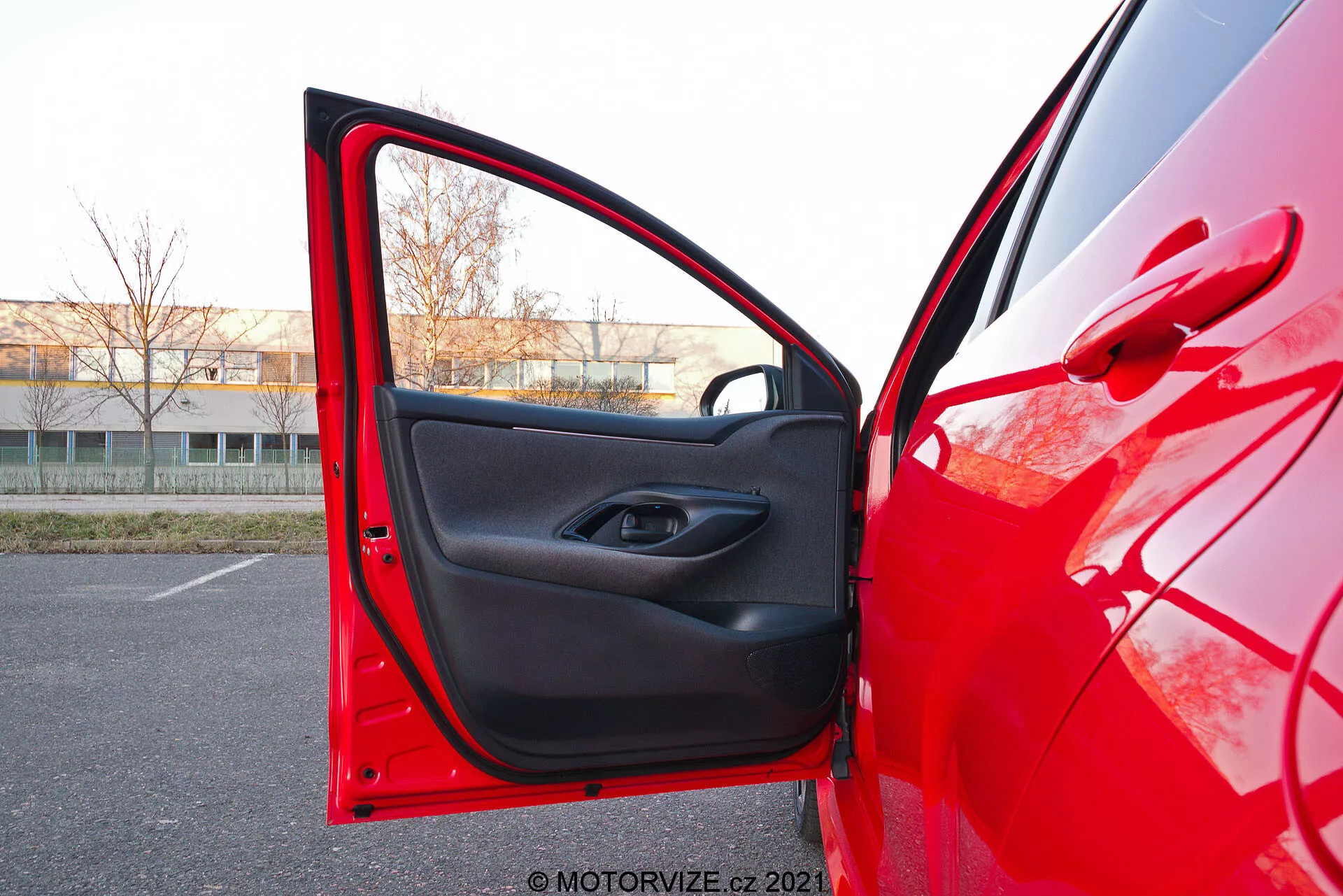 Närbild av en röd 2020 Toyota Yaris förarsida med framdörren öppen, tagen från en något låg vinkel. Kameran är placerad utanför, nära dörren, och pekar mot interiören. Den visar den ljusröda exteriörfärgen som reflekterar ljuset och den svarta interiören med en minimalistisk design. Dörren har en elektrisk fönsterknapp i handtagsfördjupningen och en förvaringsficka nedanför.
